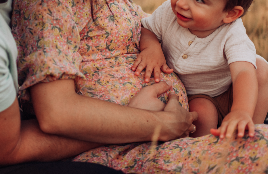 A young boy sitting on someone's lap, reaching out to touch a pregnant woman's belly, who is wearing a floral dress. The woman is holding her belly and the scene is outdoors.