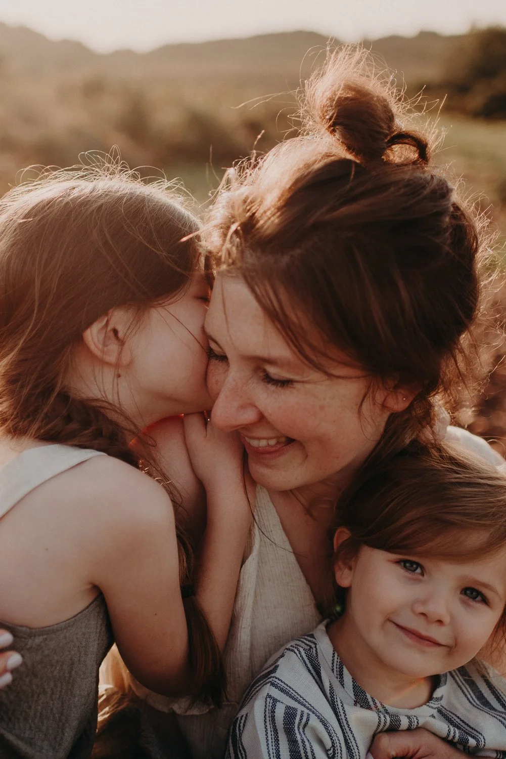 Mother and two children sharing a happy moment outdoors during sunset, with one child kissing mother on the cheek and the other smiling at the camera.
