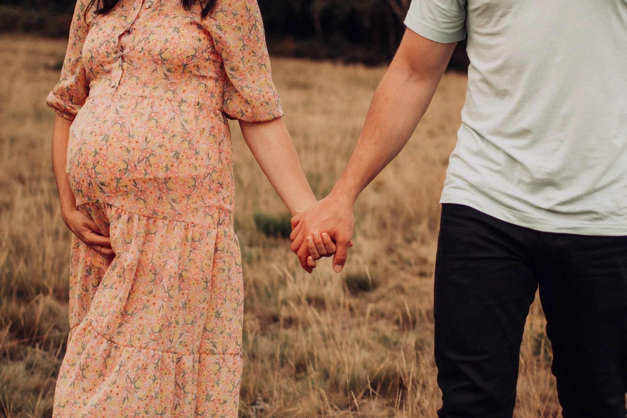 A pregnant woman in a long floral dress holding her belly with one hand, holding hands with a man in a white t-shirt and black pants, standing outdoors in a grassy field.