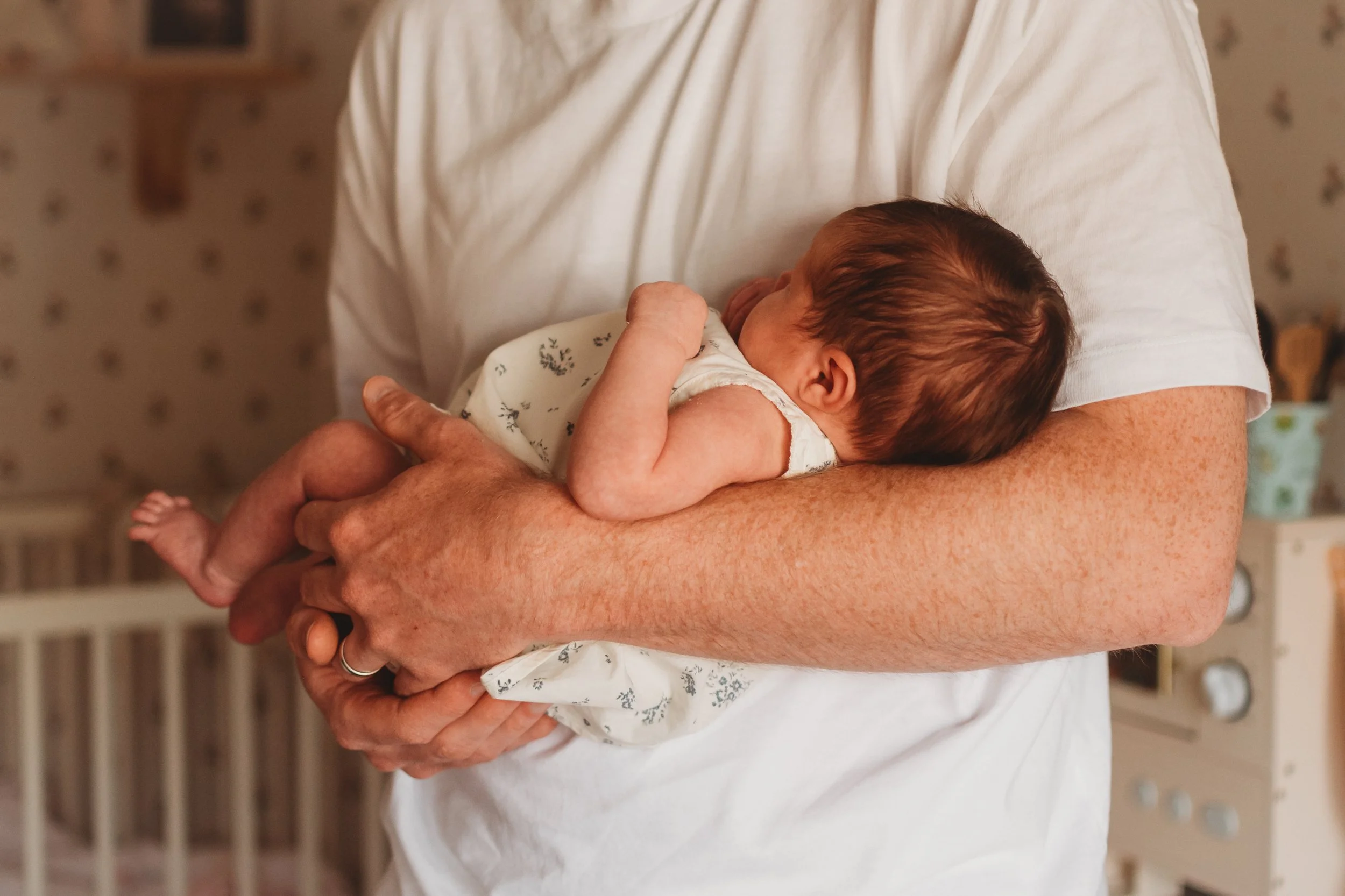An older person with freckles cradling a newborn baby in their arms, with the person's arm visible in a short-sleeved white shirt, in a cozy indoor setting. In home newborn photography in York