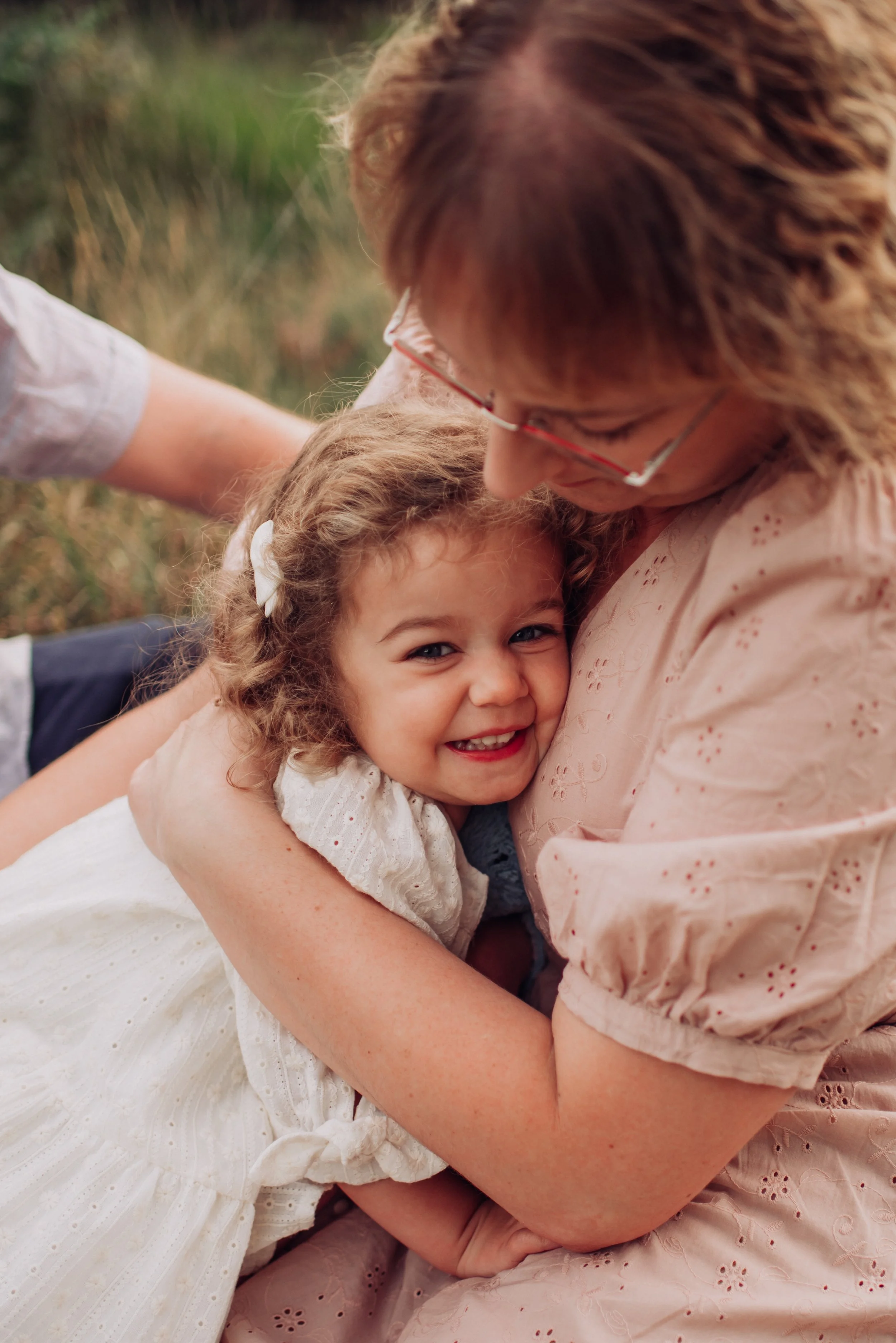 A young girl with curly hair and a white dress is happily hugging an older woman with glasses, wearing a pink embroidered blouse, outdoors in a grassy area.