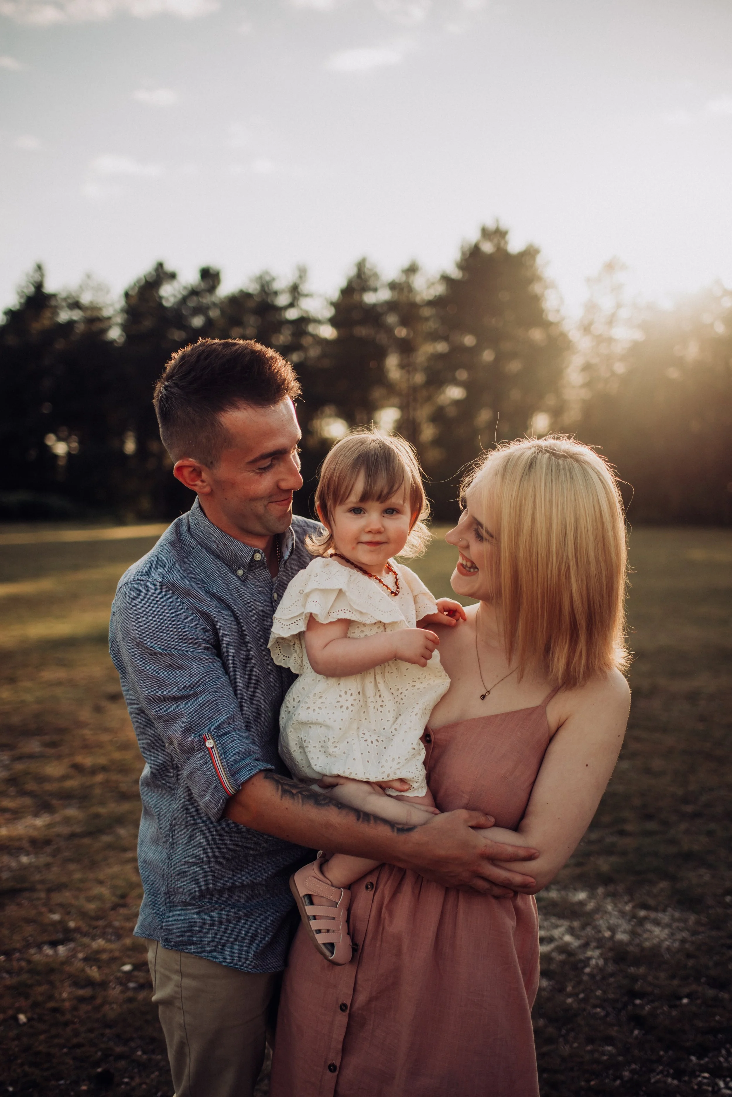 A happy family of three standing outdoors during sunset. The father, mother, and their young daughter are smiling and looking at each other. The father is holding the daughter, and the mother is smiling at her. The background shows trees and a soft, warm sky.