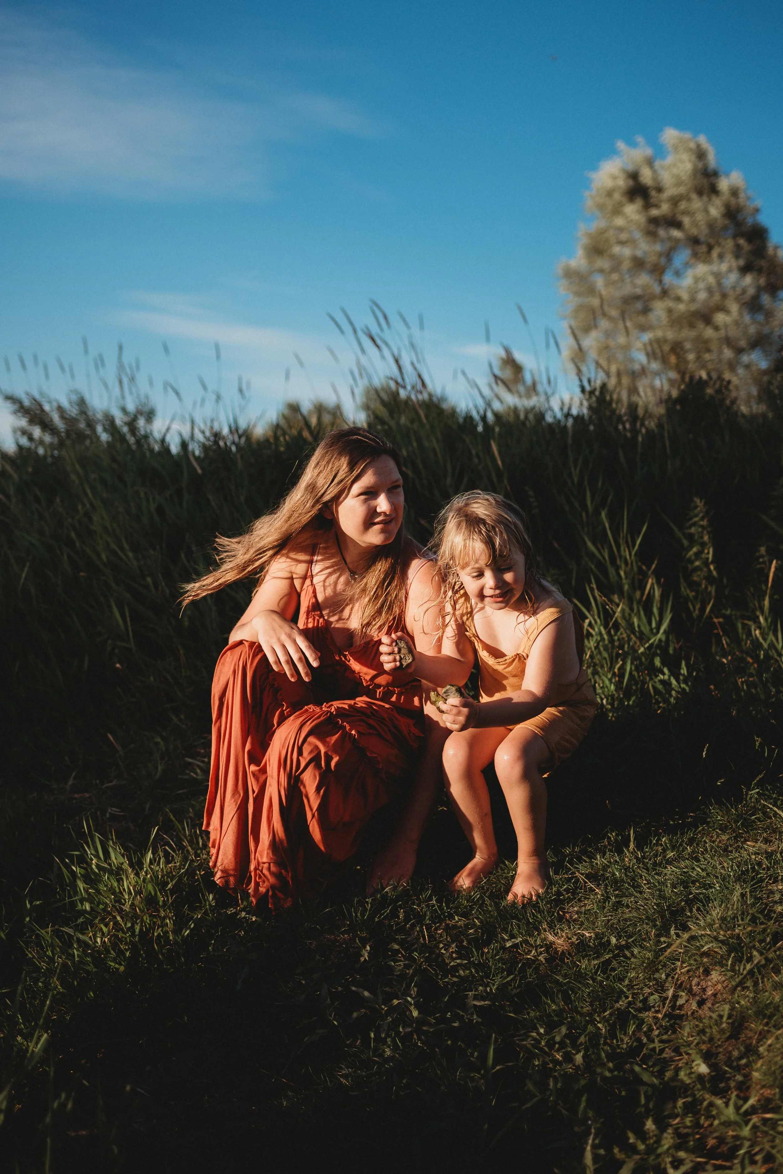A woman and a young girl sitting on grass outdoors, both holding rocks, with tall grass and a blue sky in the background.