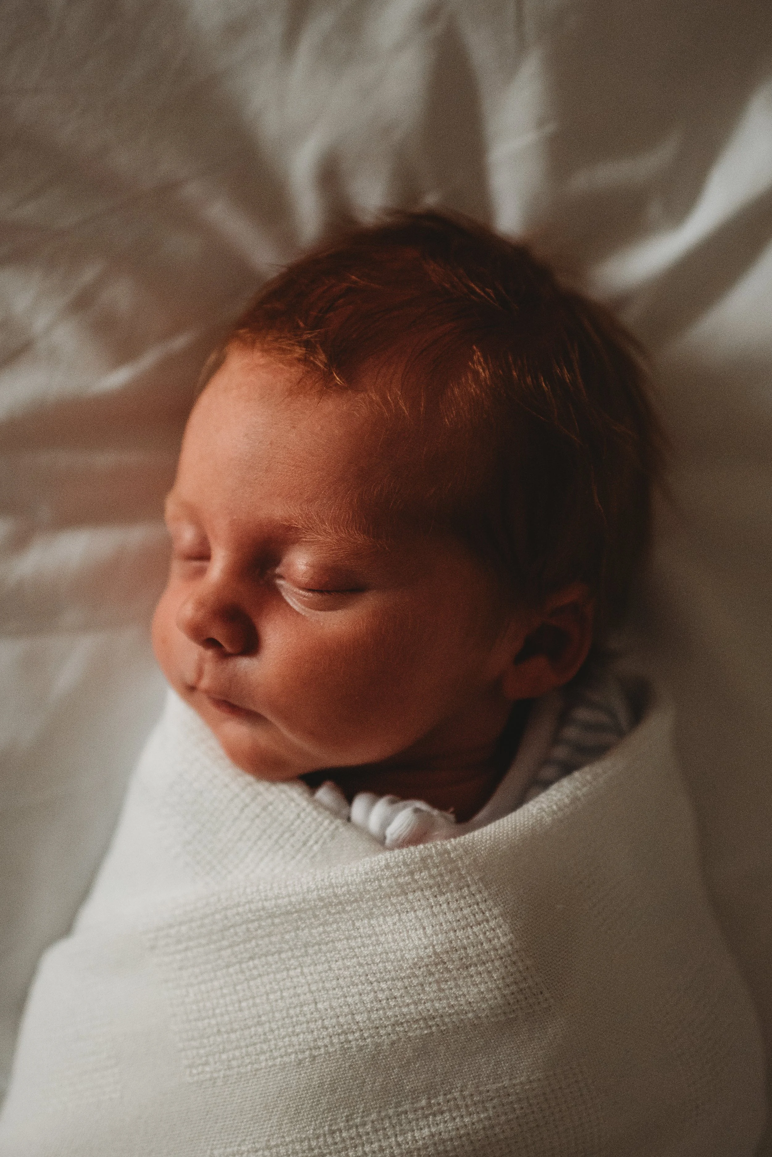 Close-up of a sleeping newborn baby wrapped in a white blanket, lying on a soft surface.