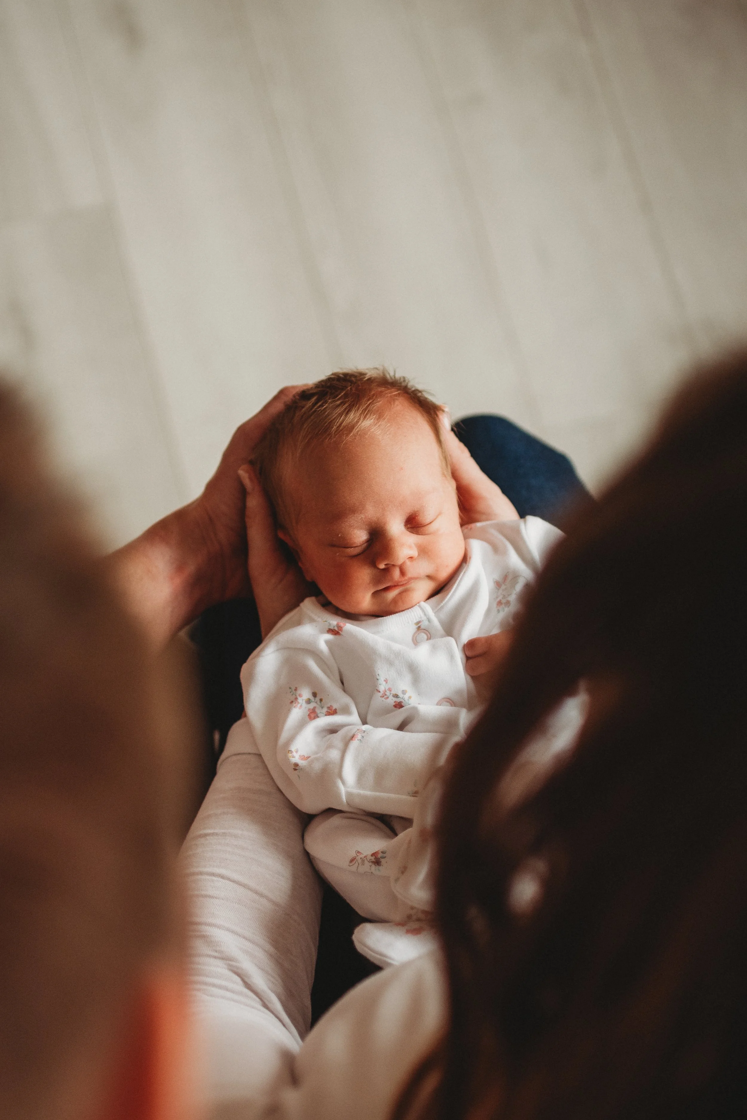 A baby with red hair sleeping in an adult's lap, with an adult's hands gently holding the baby's head.