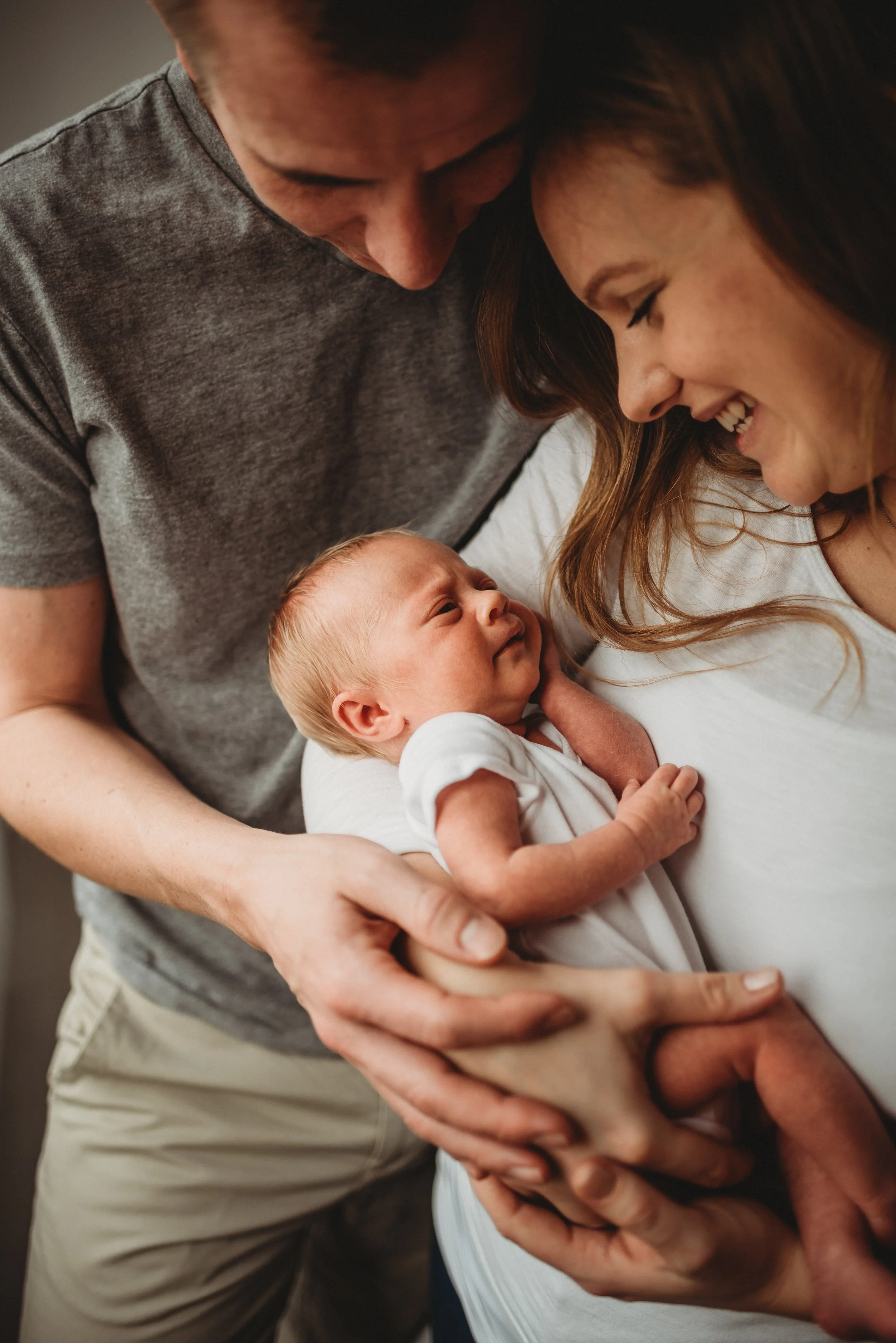 Lifestyle newborn photography of a  couple with their newborn baby, smiling and looking at each other while holding the baby in a close embrace.