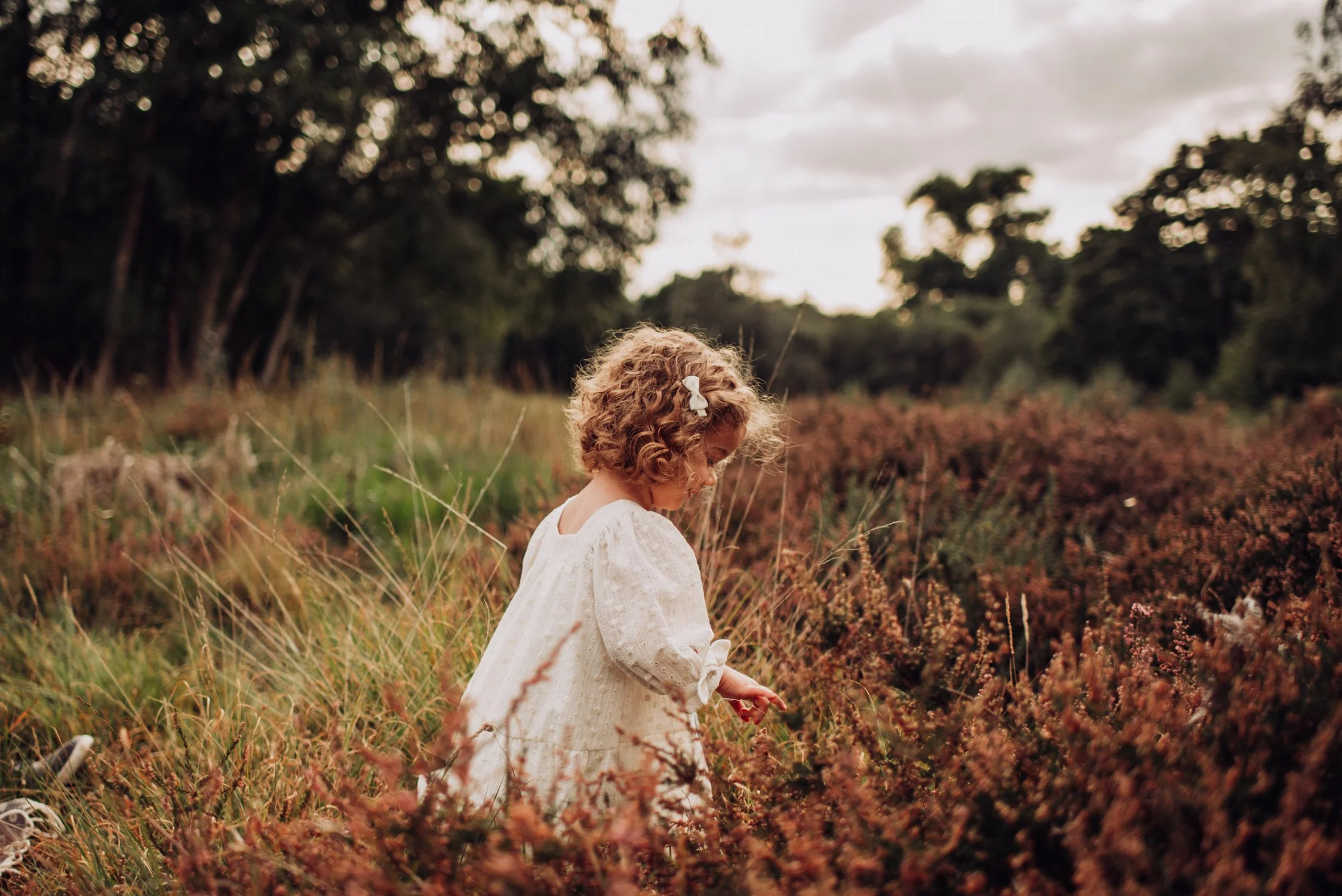 A young girl with curly hair and a white dress stands in a field of brown and green plants, with trees and a cloudy sky in the background.