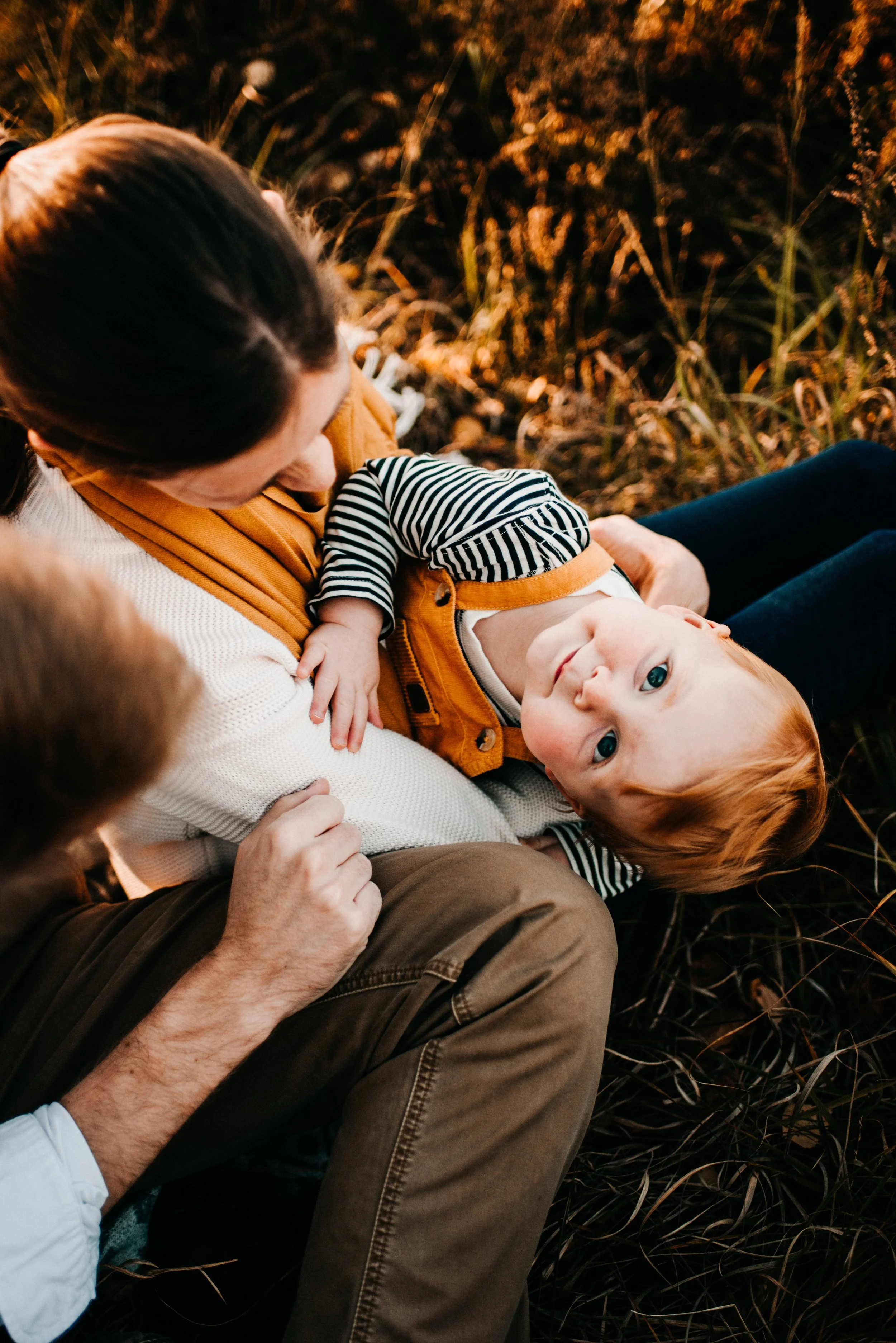 A baby with red hair lying on a woman's lap in a field of grass, looking up at the camera while another person, possibly a man, touches the baby's hand.
