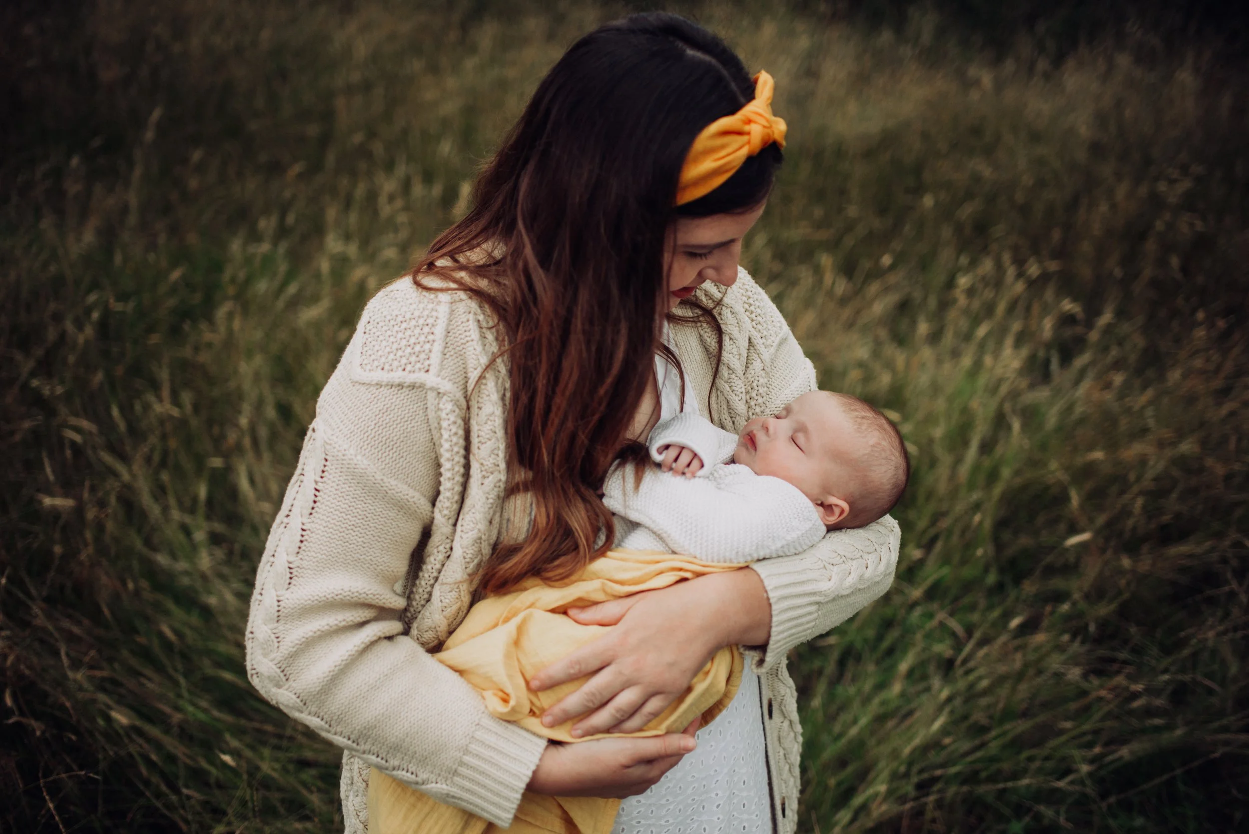 A woman with long brown hair and a yellow headband holding a sleeping baby wrapped in a yellow blanket in a field of tall grass.