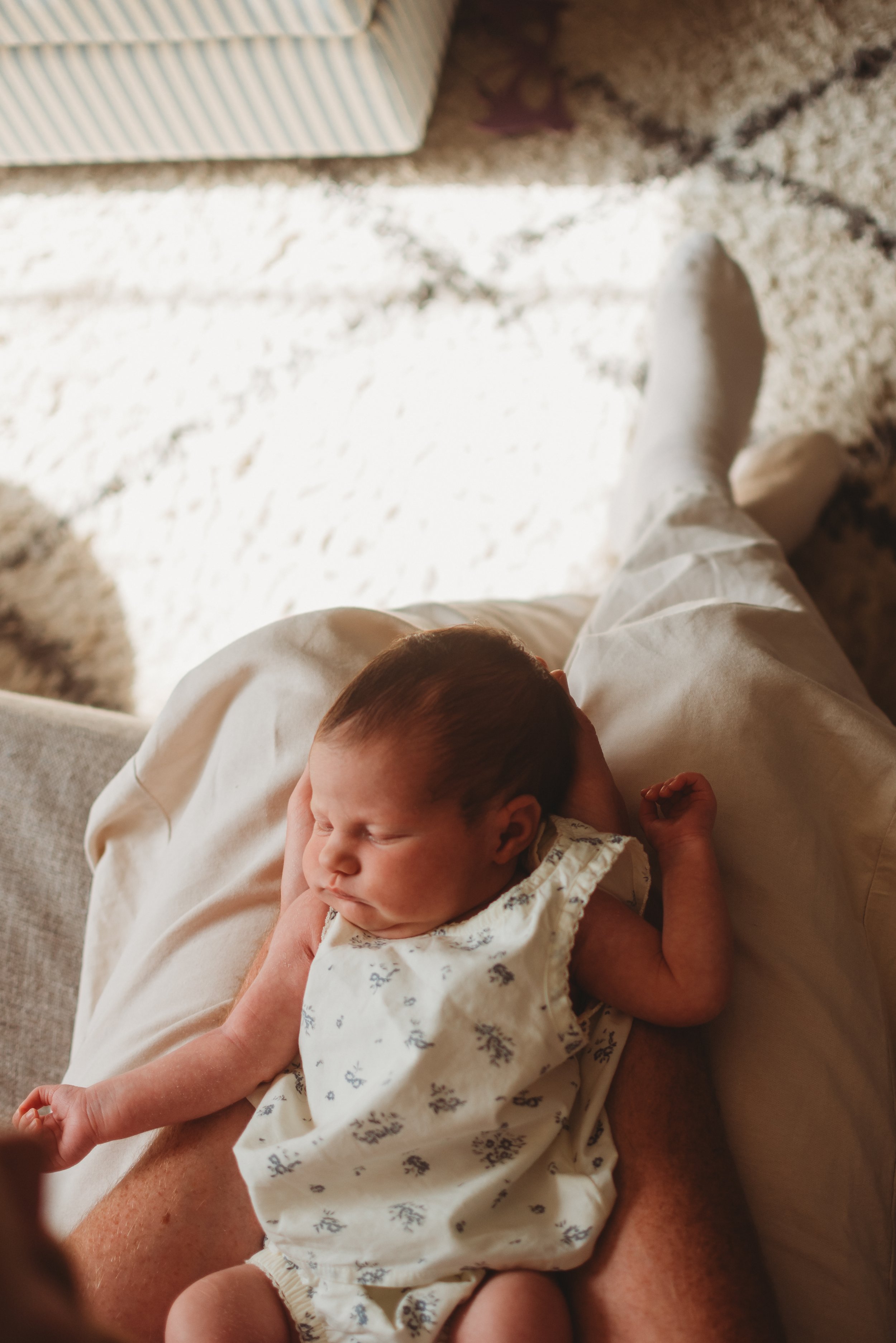 Close-up of a person holding a sleeping baby on their lap, with the baby wearing a white floral dress and the adult wearing light-colored pants, in a cozy indoor setting. In home newborn photography in York