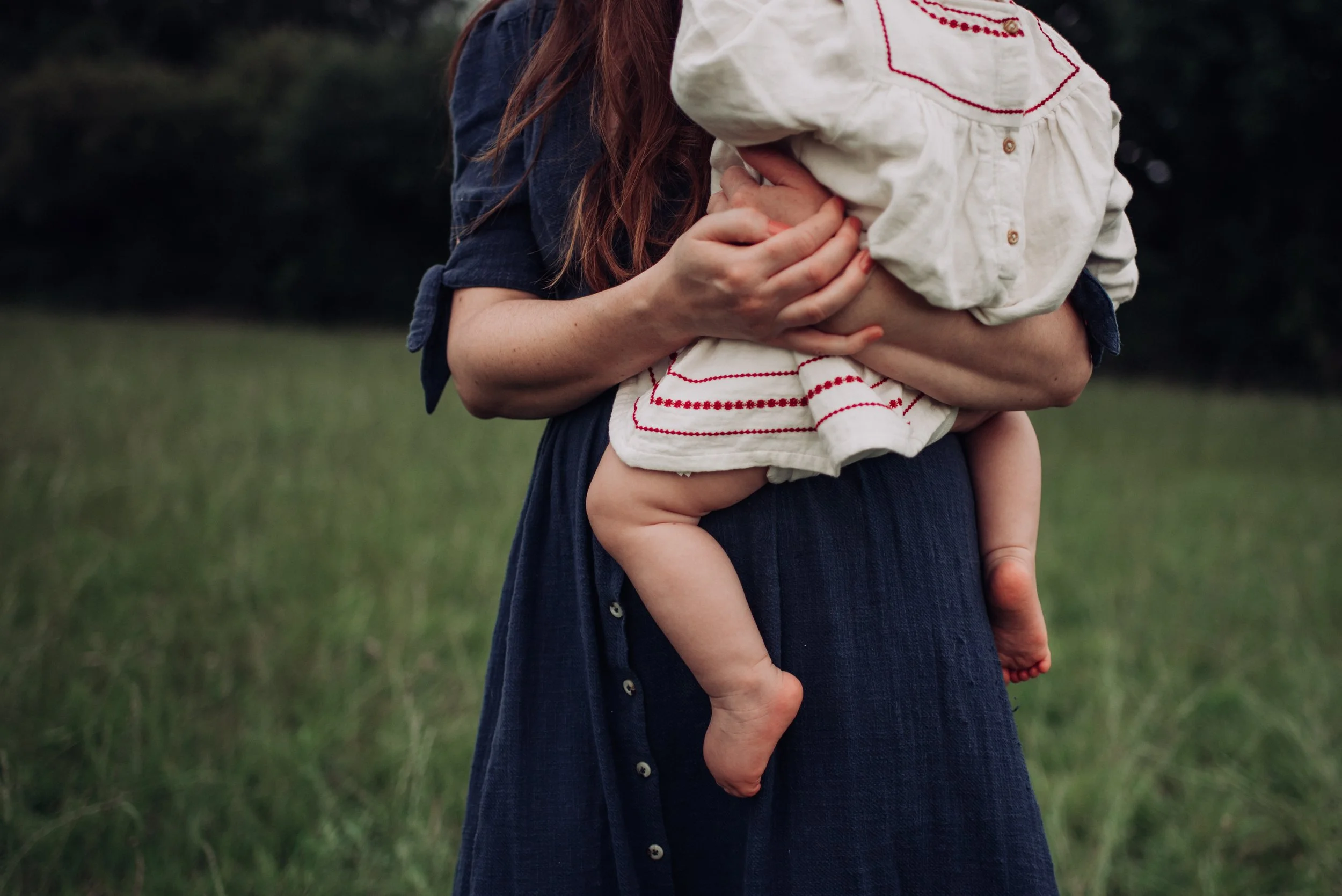 A woman holding a young child outdoors in a grassy area, with part of her red hair and dark blue dress visible.