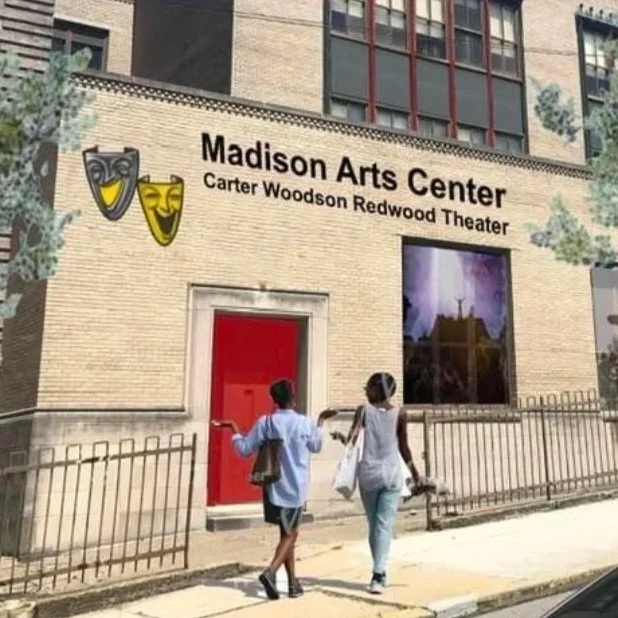 Two women walking past the Madison Arts Center, located in the Carter Woodson Redwood Theater. The building has a brick facade with a large red door, theatrical masks on the wall, and a big window displaying a scene or poster. The women are carrying bags and walking toward the theater entrance.