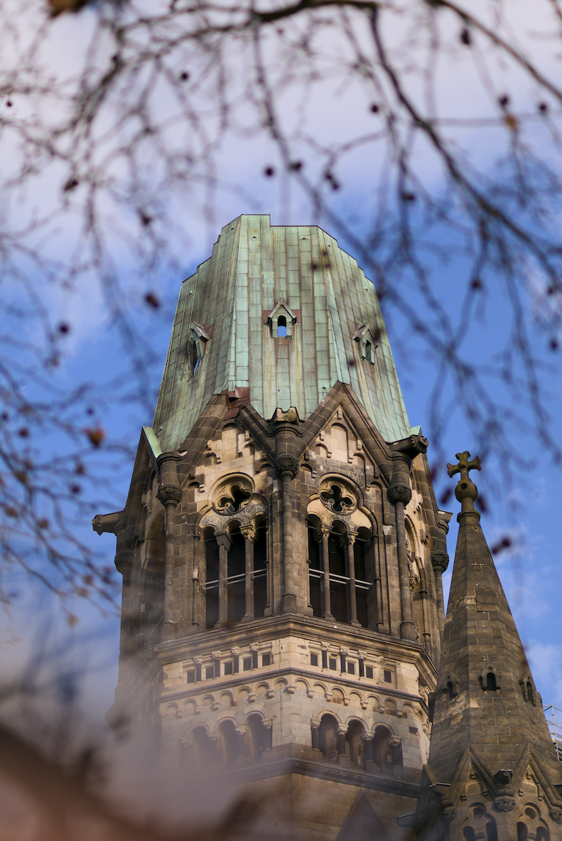 Der Blick nach oben auf einen gotischen Kirchturm, eingerahmt von Baumzweigen und -ästen im Vordergrund, mit blauer Himmel im Hintergrund.