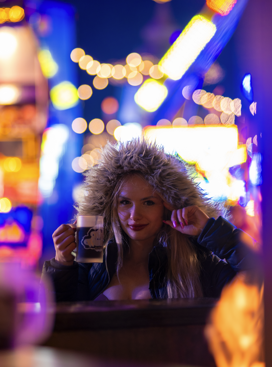 Junge Frau mit warmer Kapuze, die einen Drink hält und in einer festlich beleuchteten Stadt bei Nacht ist.