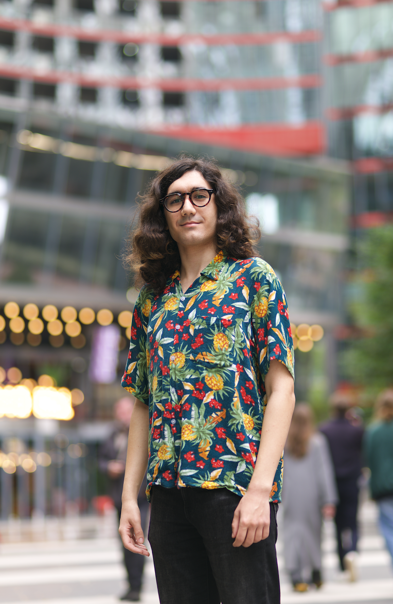 Junge mit langem, lockigem Haar und Brille steht in einer urbanen Umgebung mit moderner Architektur und Menschen im Hintergrund.