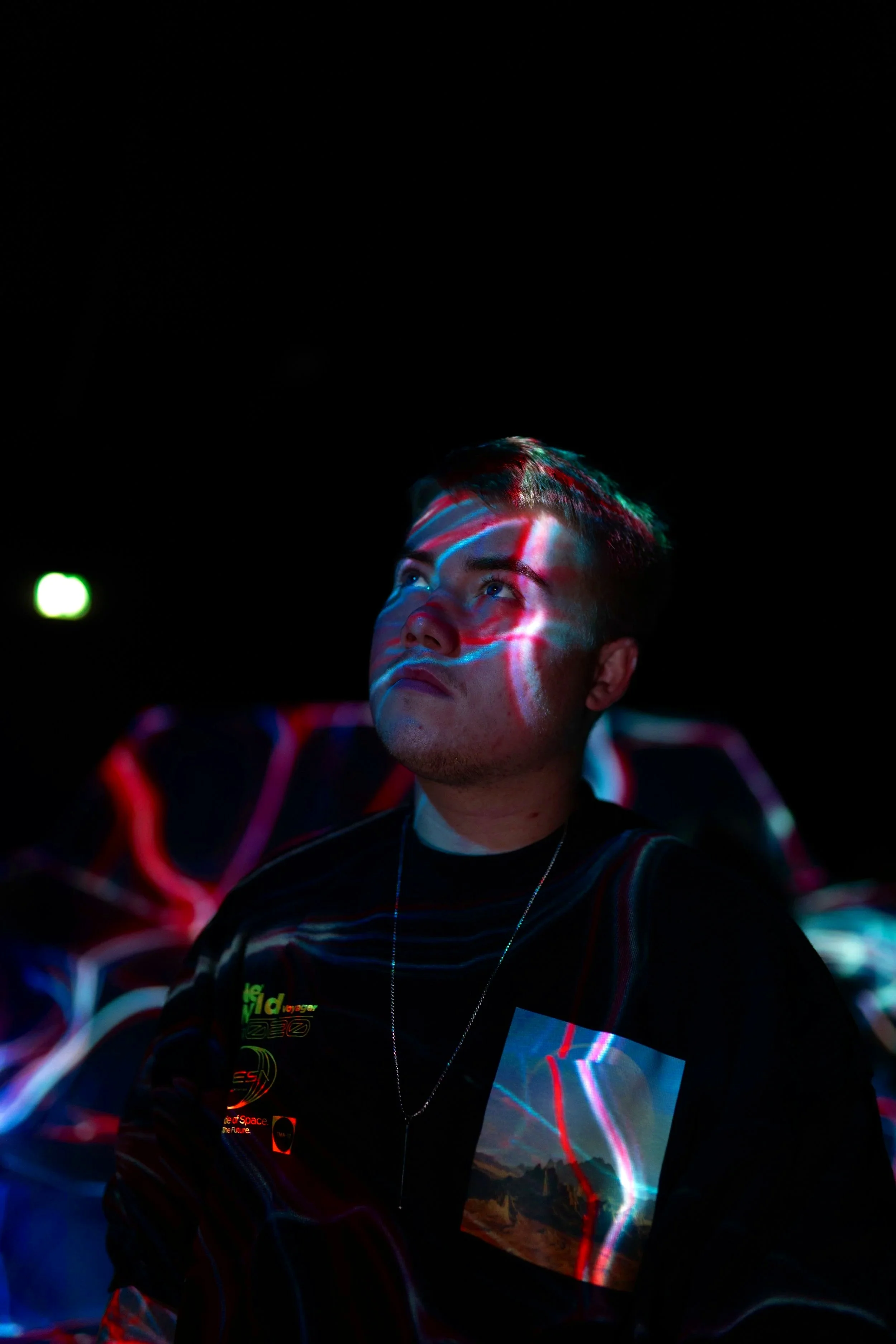 Junge Person sitzt in einem dunklen Raum, beleuchtet von bunten Lichtmustern auf Gesicht und Kleidung.