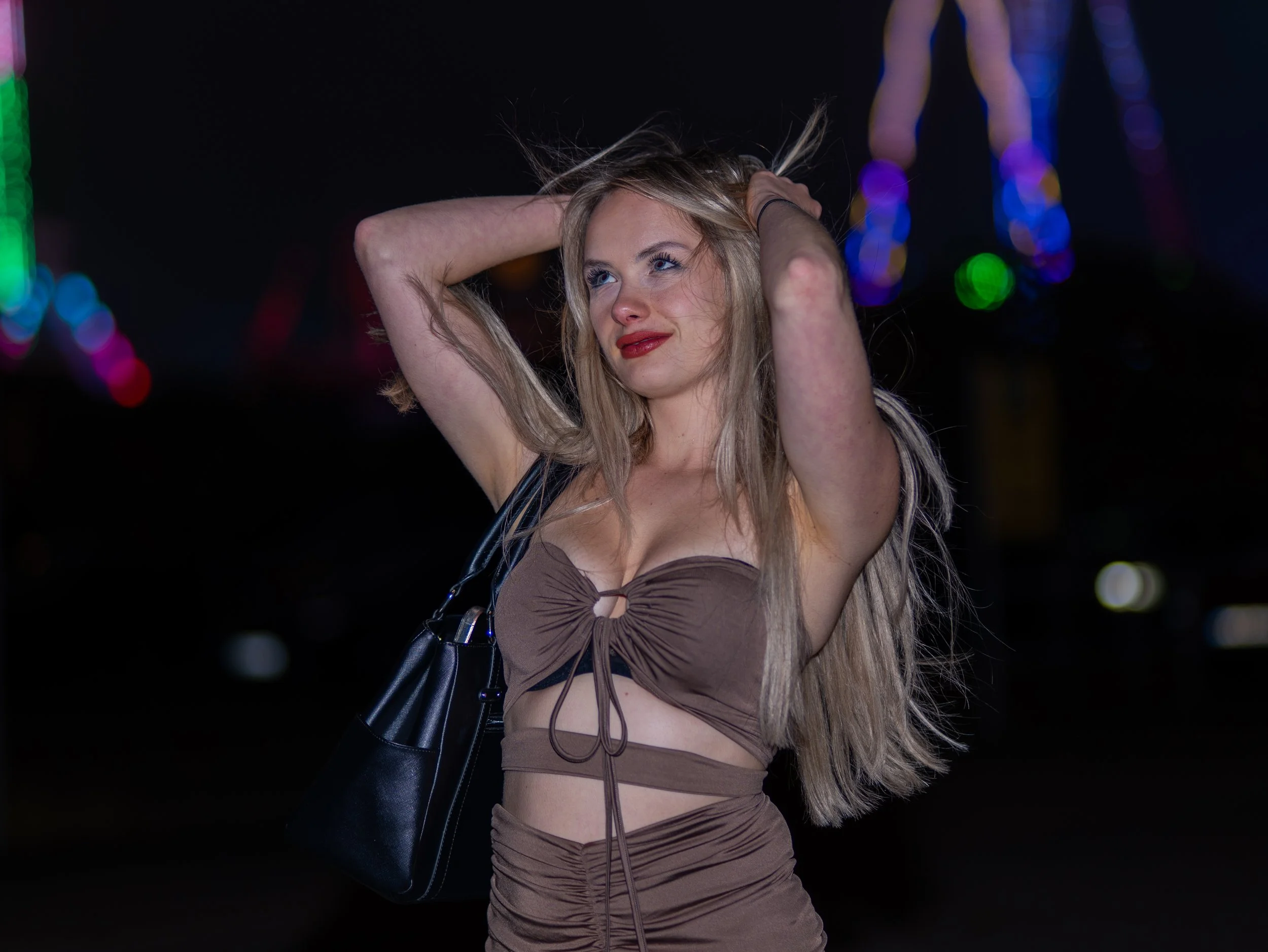 Junge Frau mit langer blonder Haar, trägt ein braunes Kleid und trägt eine schwarze Tasche, steht vor bunten Lichtern bei Nacht.