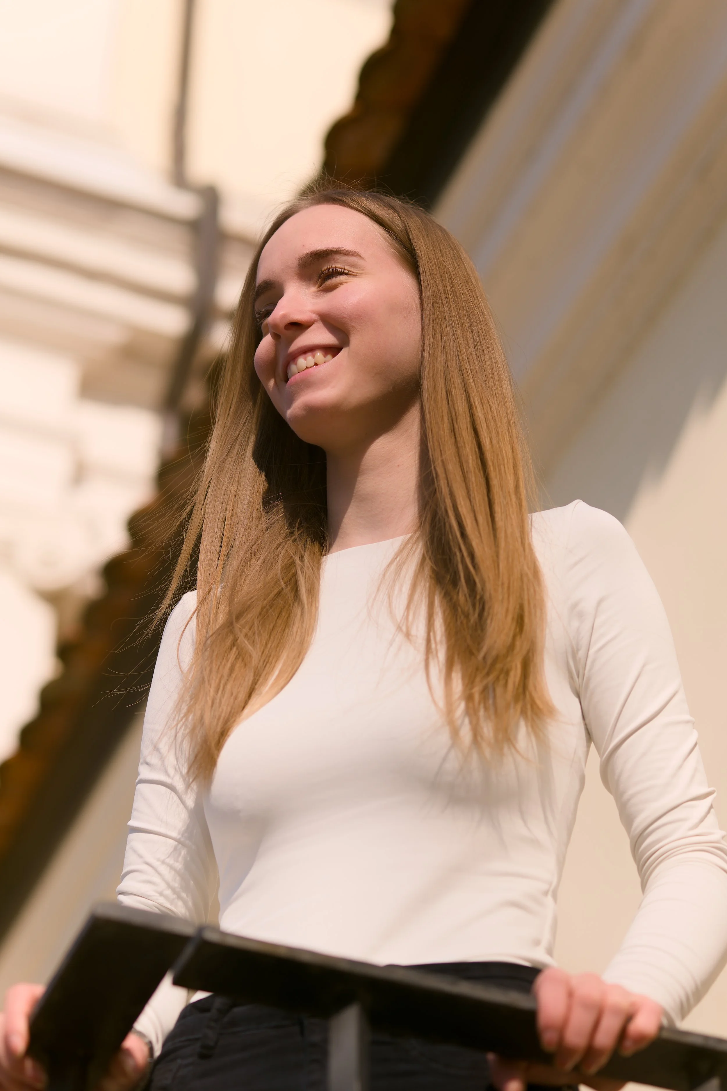Junge Frau mit langen Haaren in weißem Oberteil, lächelnd und im Sonnenlicht, vor einem Gebäude.
