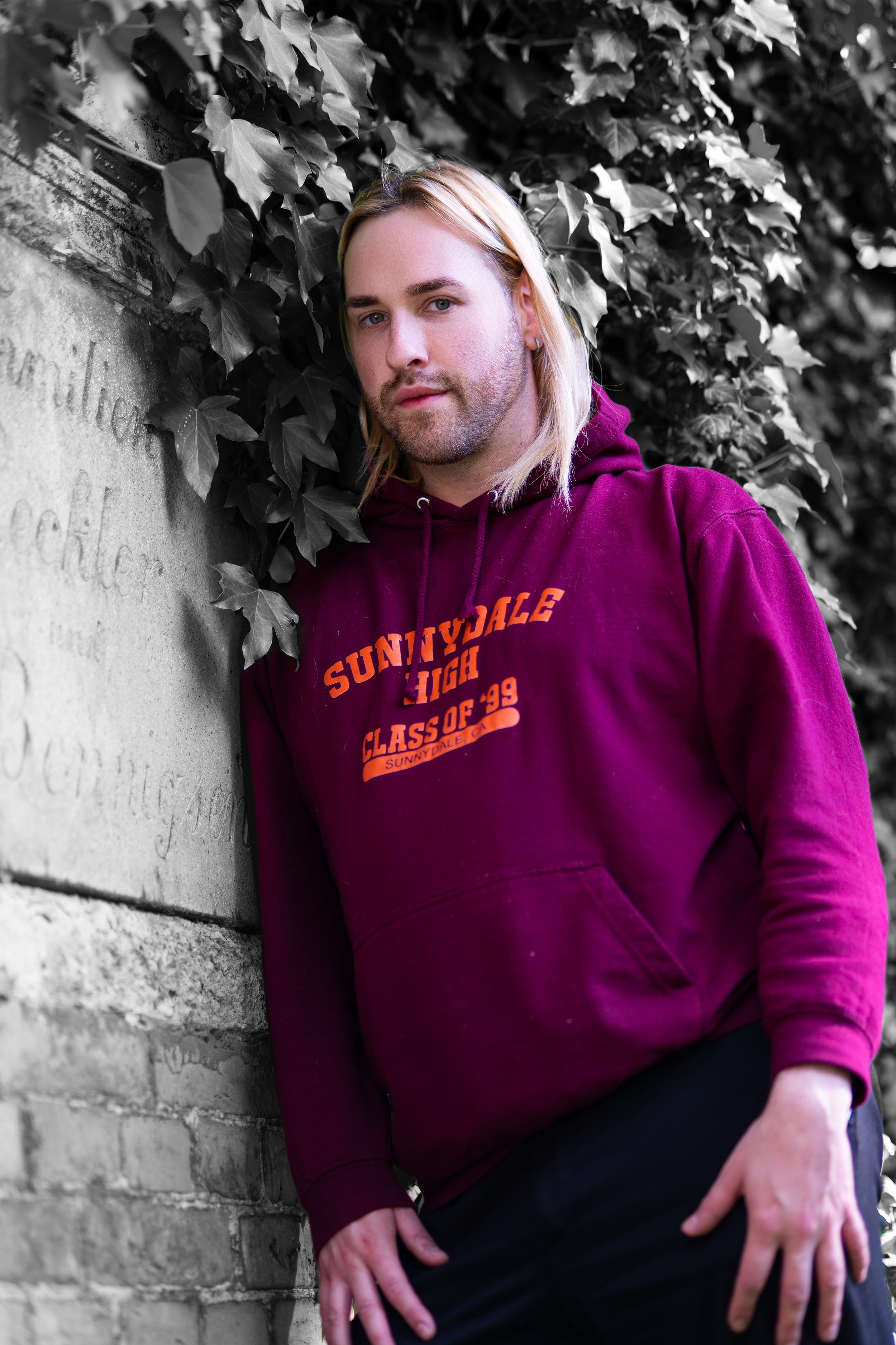 Junge Person mit blonden Haaren in einer burgunderfarbenen Kapuzenjacke, die an einer Wand mit grünen Blättern lehnt.