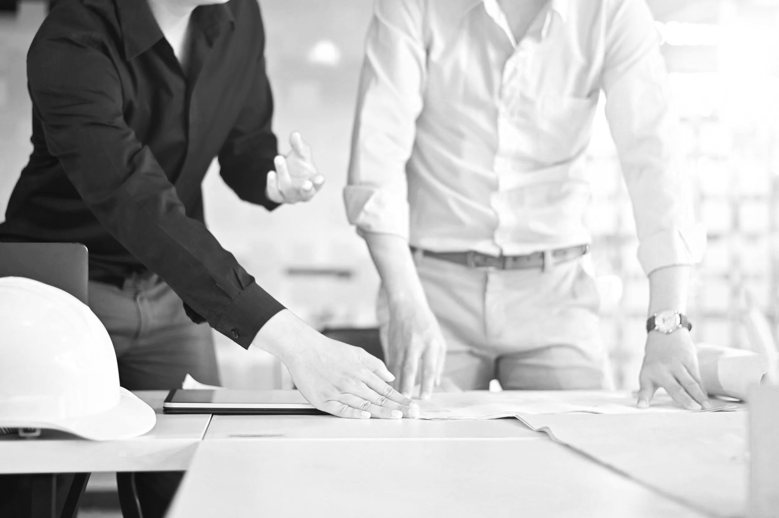Two individuals in business attire collaborating over architectural plans at a table, with a construction hard hat placed nearby.