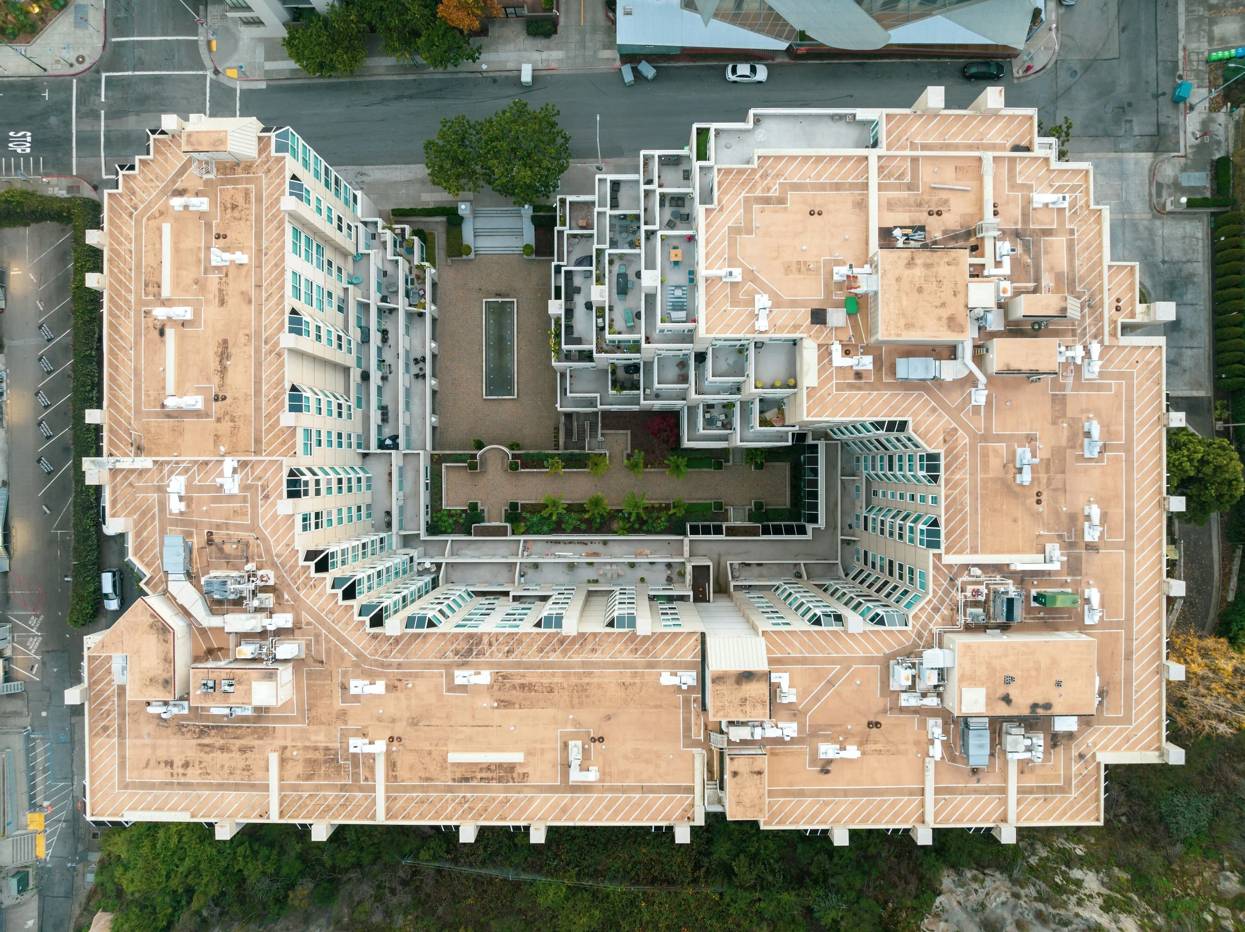 Bird's eye view of a large multi-story apartment building with rooftop amenities, surrounding streets, parking lot, and trees.