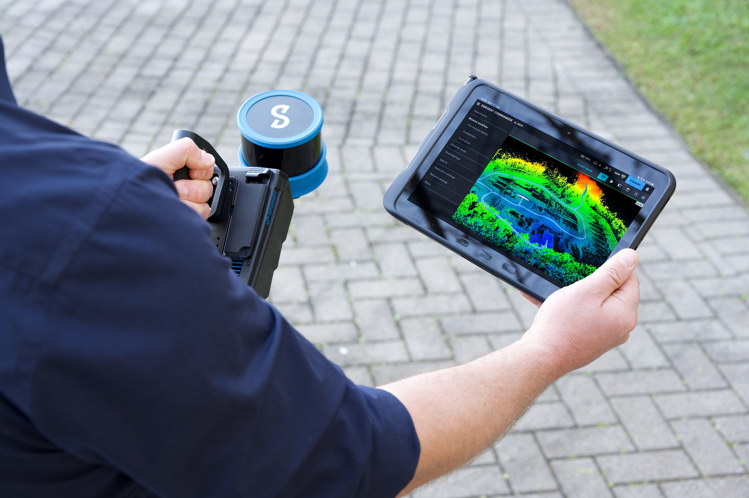 Person holding a tablet displaying a LiDAR scan of a parking lot with green, yellow, and red color mapping; also holding a handheld device with a scanning sensor.