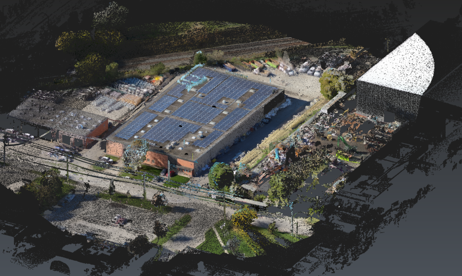 Aerial view of a commercial or industrial building with solar panels on the roof, surrounded by parking lots, trees, and nearby structures.