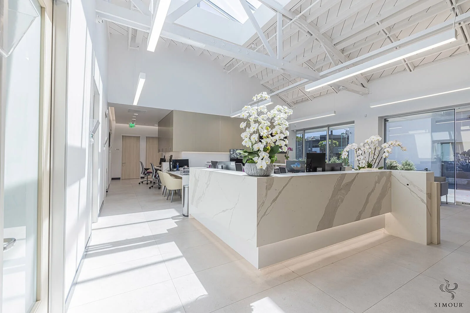 Modern office reception area with white marble desk and large floral arrangements, bright natural light, and glass sliding doors.