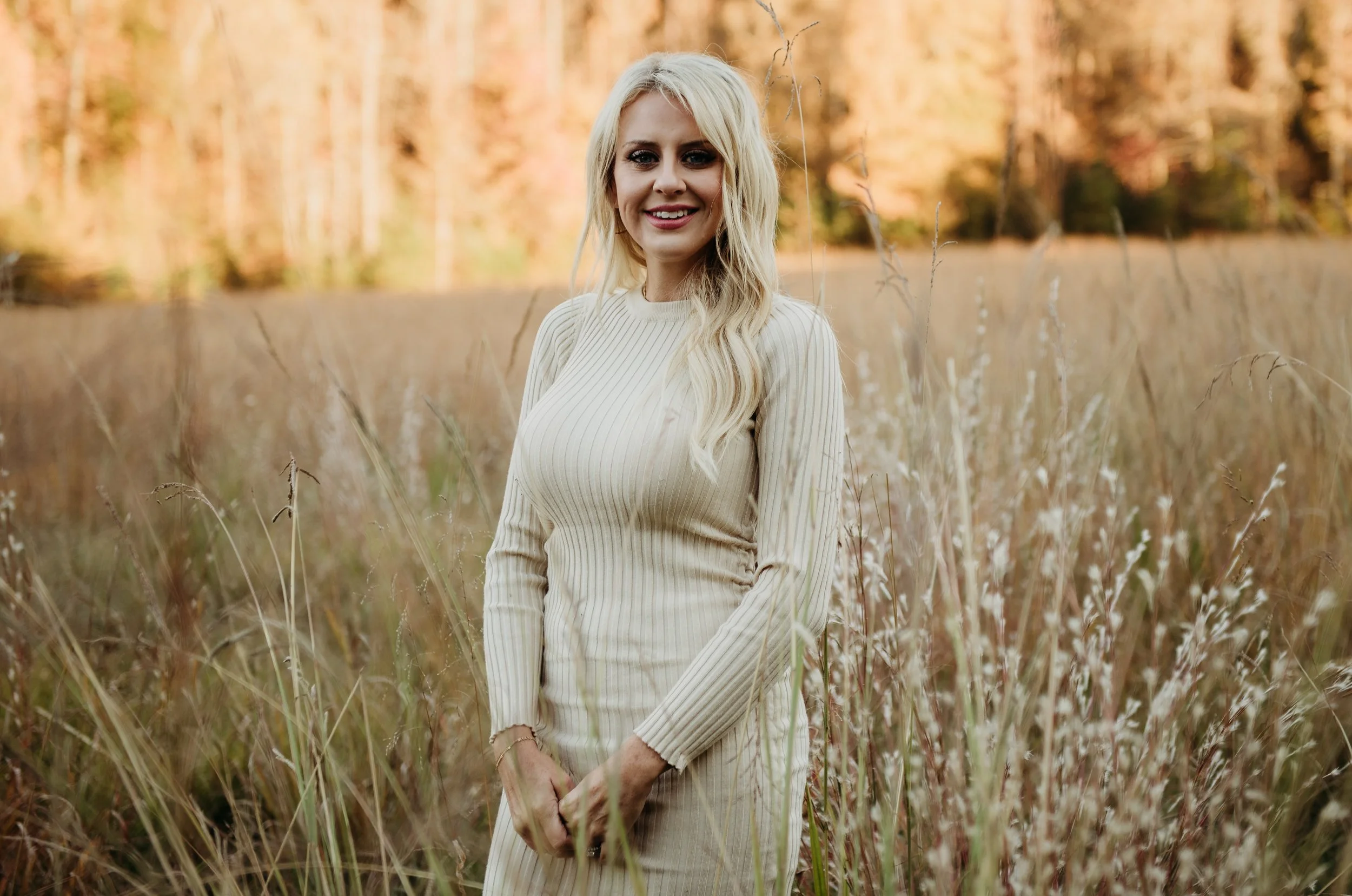 A young woman with long blonde hair smiling in a field of tall grass during autumn with trees in the background.