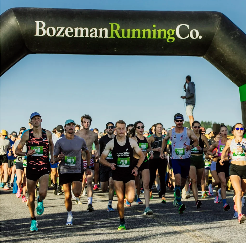 Runners start a race under an archway that says 'Bozeman Running Co.' on a sunny day during the Bozeman Half Marathon.