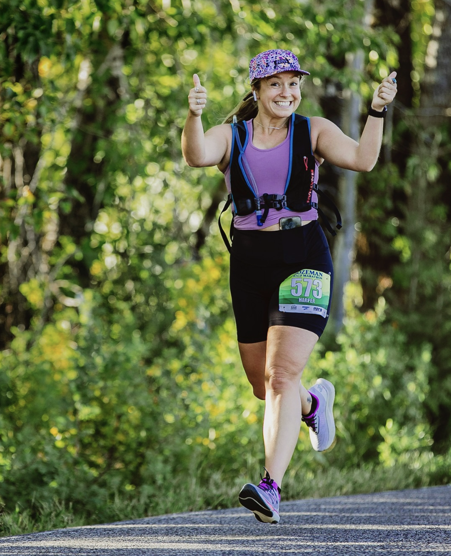Female marathon runner smiling and giving thumbs up while running on a forest trail, wearing a purple tank top, black shorts, colorful running shoes, and a floral cap.