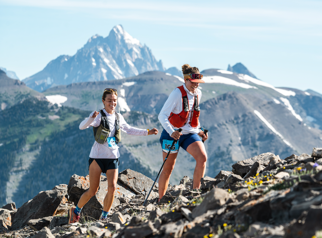 Two women trail running on a rocky mountain trail with snow-capped peaks and a blue sky in the background at Jackson Hole Mountain Resort.