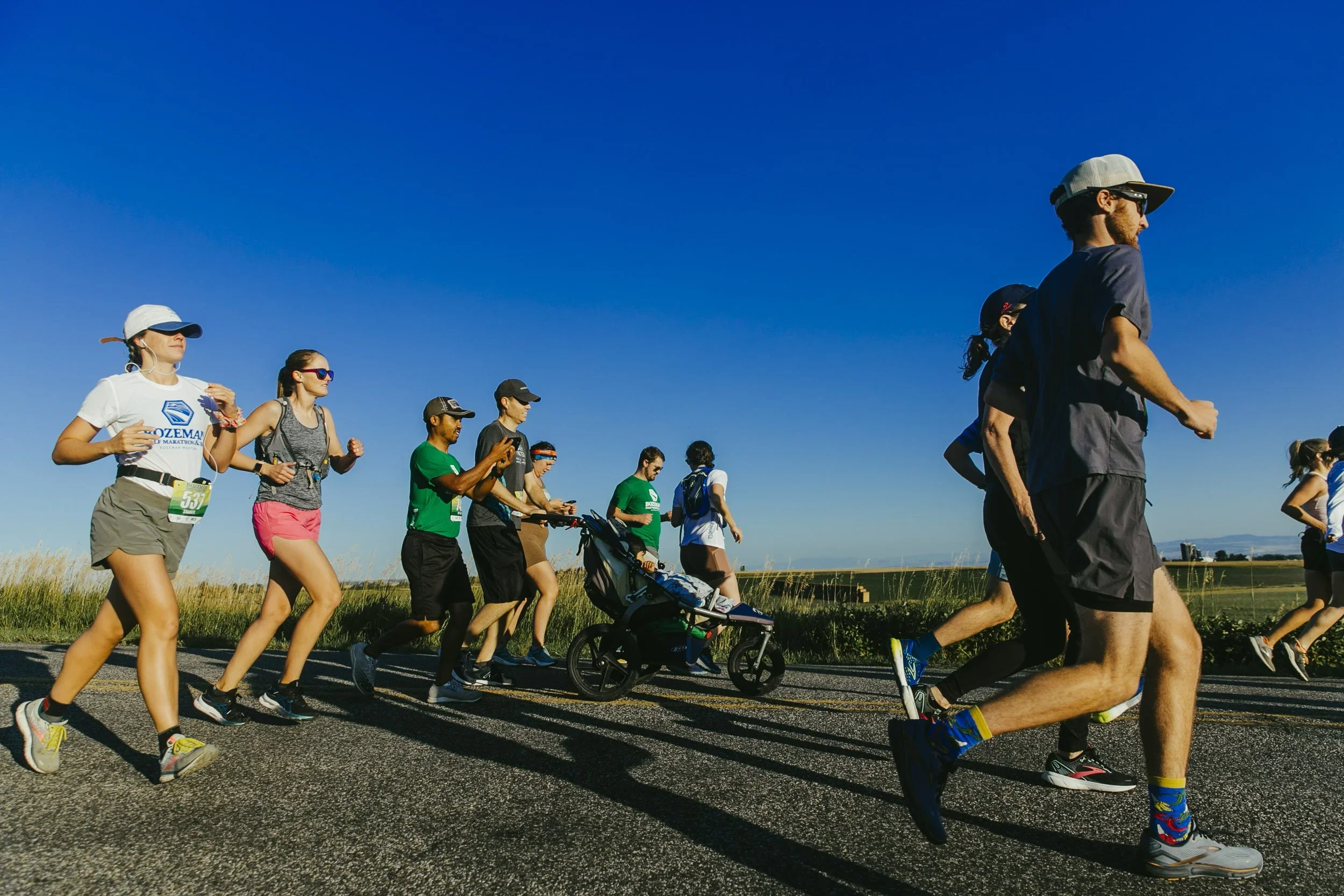 Group of people running in a race on a sunny day, some wearing athletic gear and pushing a stroller, with open fields and clear blue sky in the background.