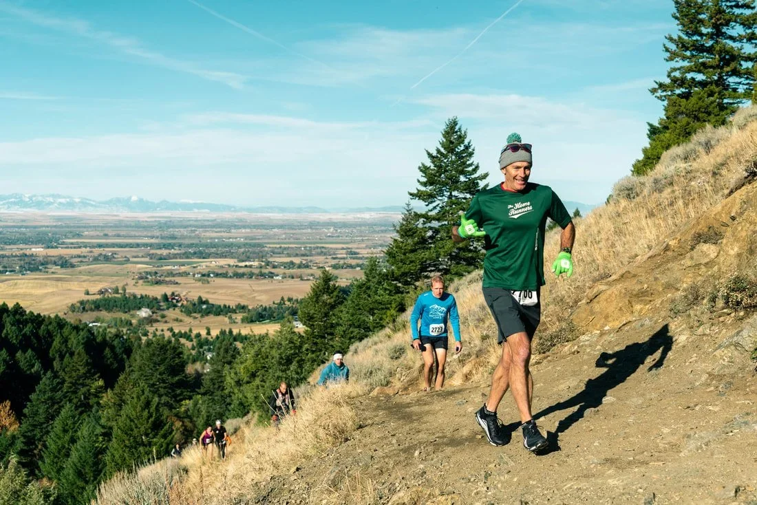 People trail running up a mountain with a scenic valley view and snow-capped mountains in the distance.