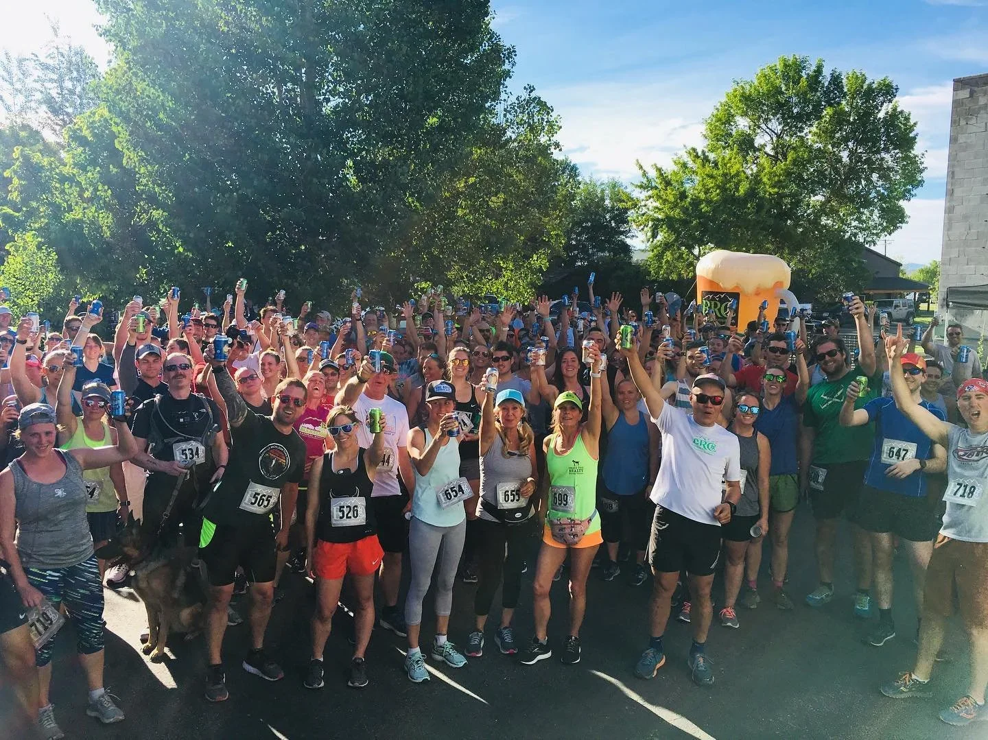 Large group of people participating in an outdoor event, many holding up cans and smiling at the camera, with trees and a blue sky in the background.