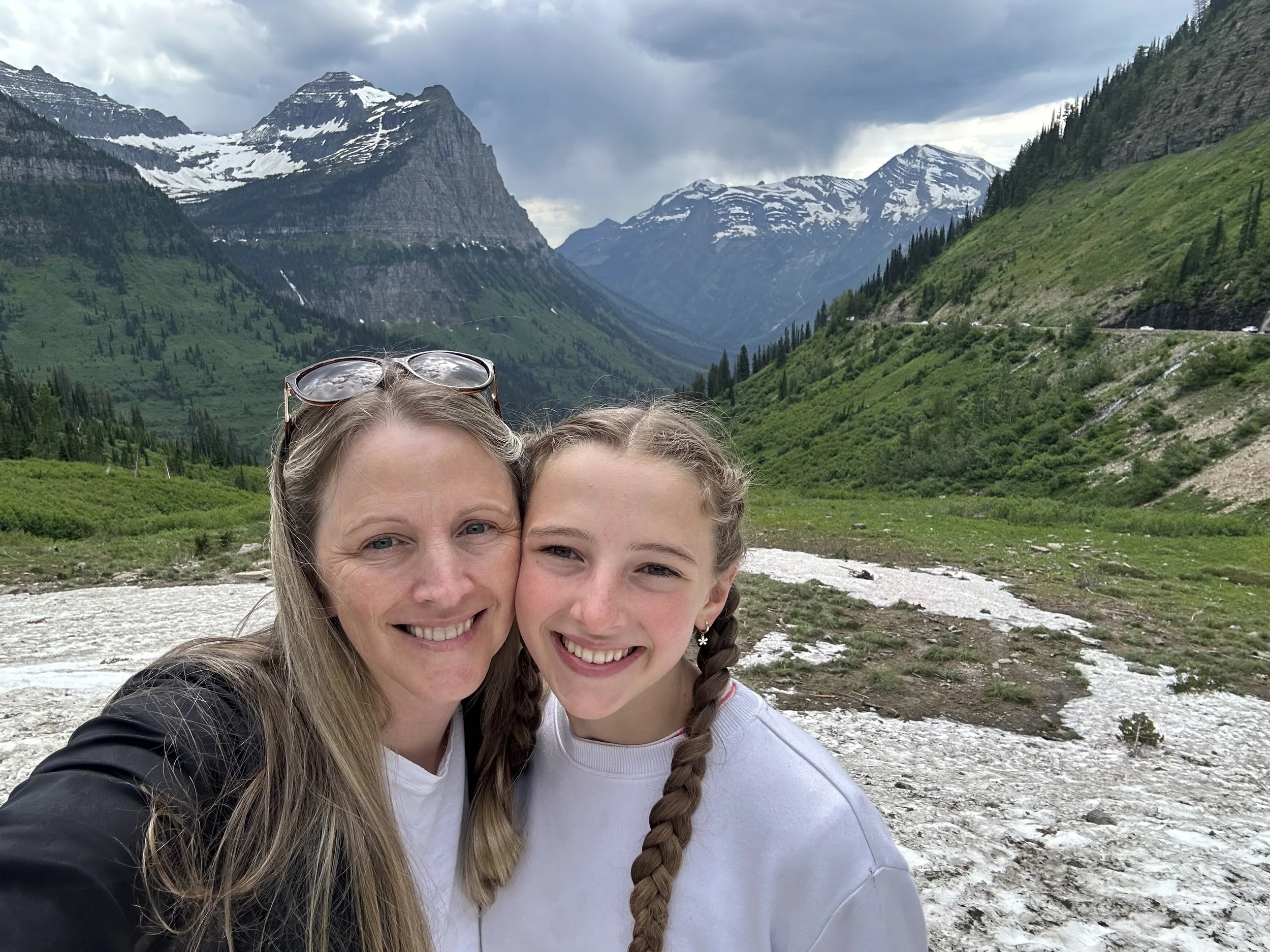 Two women smiling and hugging, standing on a rocky riverbank with lush green mountains and snow-capped peaks in the background.