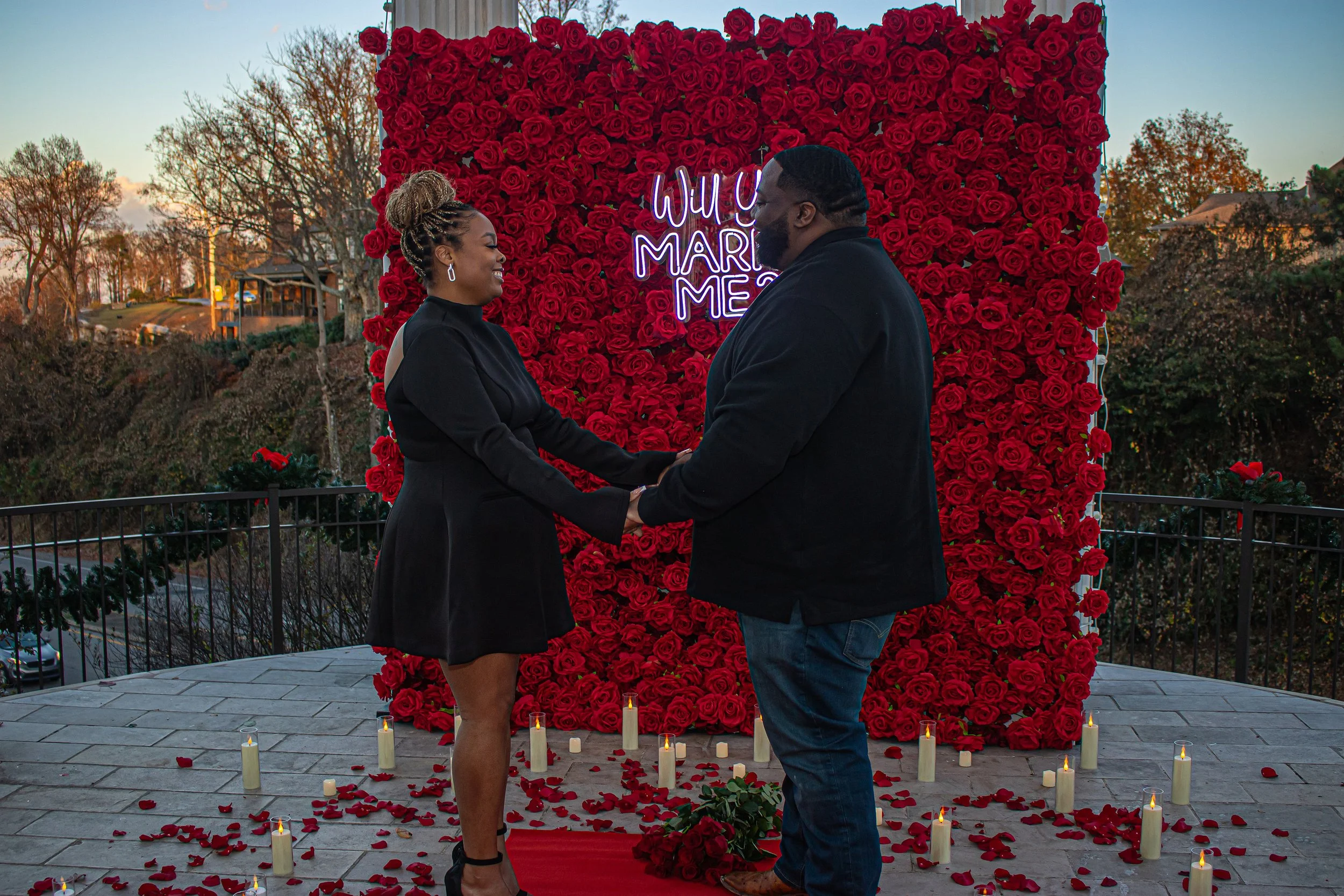 A couple holding hands and smiling at each other during a marriage proposal ceremony on a balcony with a floral backdrop of red roses and a neon sign that says "Will You Marry Me." Candles and rose petals are on the floor around them.