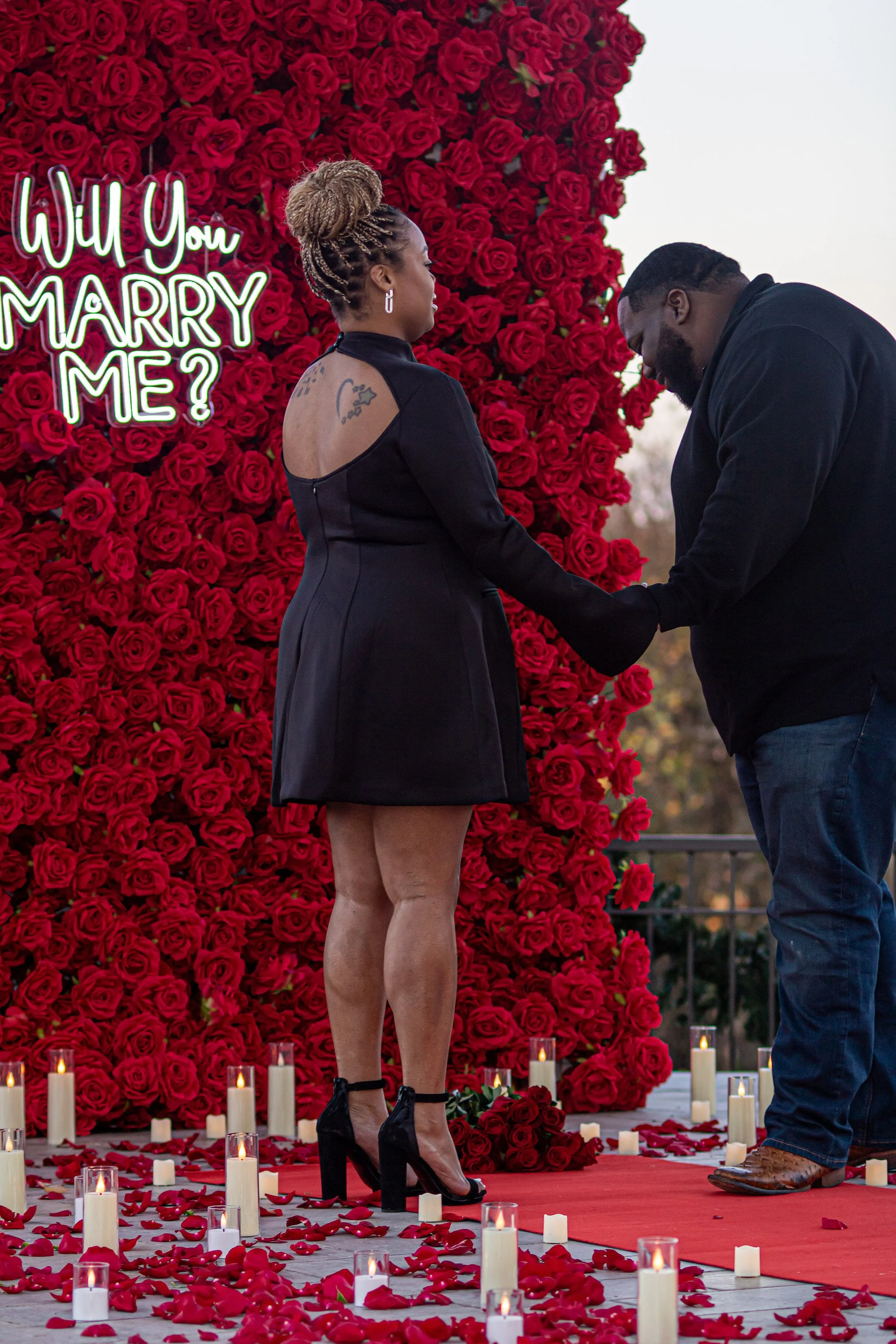 A man is proposing marriage to a woman during a romantic setup decorated with red roses, candles, and rose petals, with a neon sign that reads 'Will You Marry Me?'
