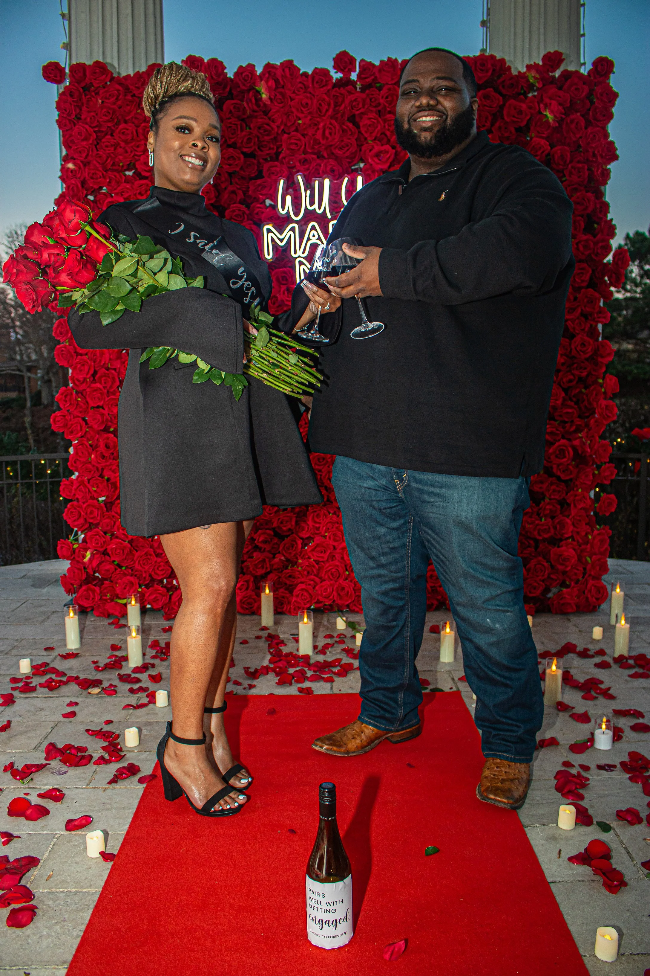 A woman in a graduation gown holding a bouquet of red roses stands on a red carpet next to a man holding a wine glass. They are outdoors in front of a backdrop made of red roses with a neon sign that says 'Will you marry me?'. There are candles and r