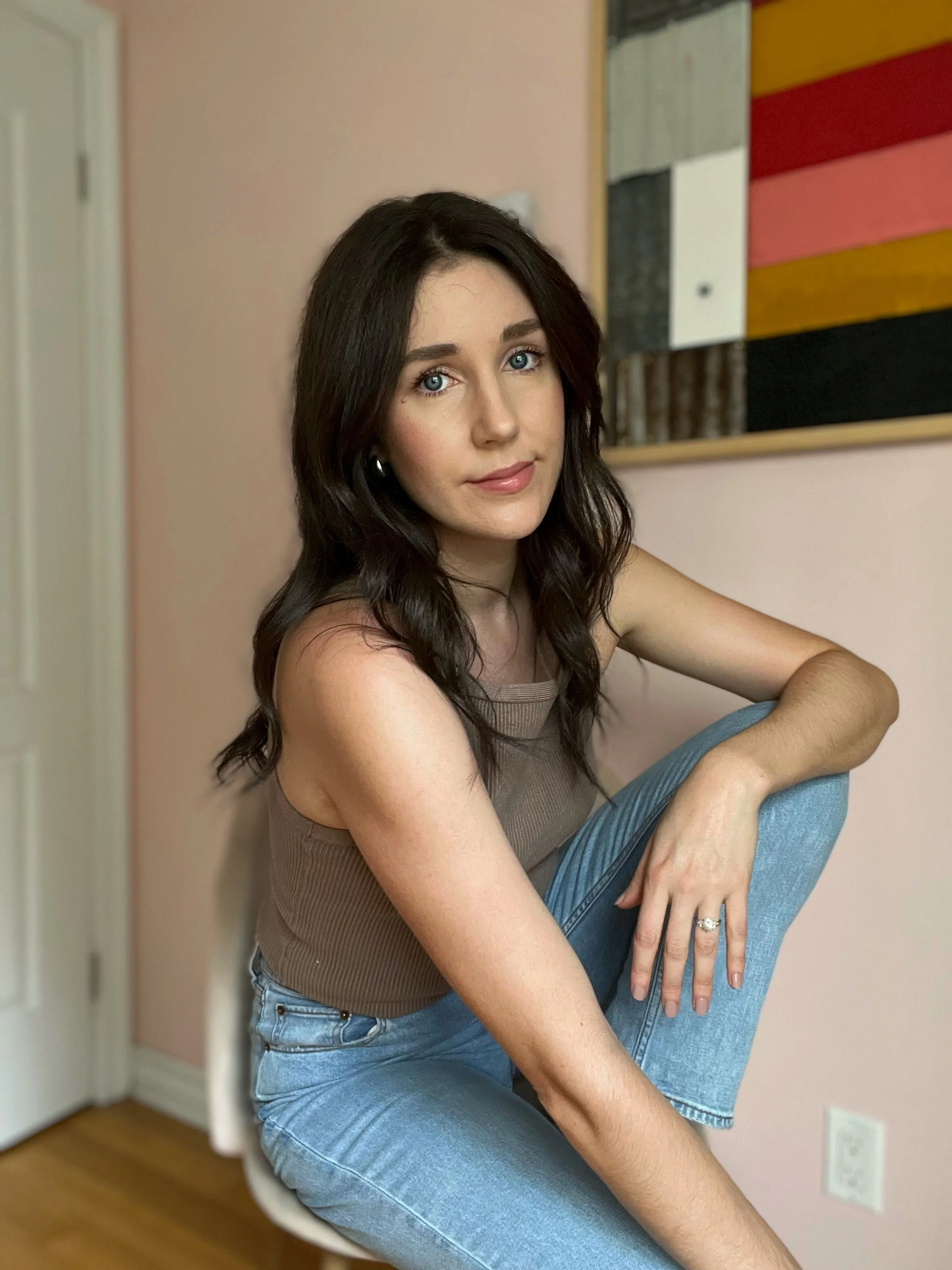 Portrait of Madeleine Gagné. She has long dark hair, blue eyes, wearing a sleeveless brown top and light blue jeans, sitting on a white chair in a room with pink walls and a colourful abstract painting hanging behind her.