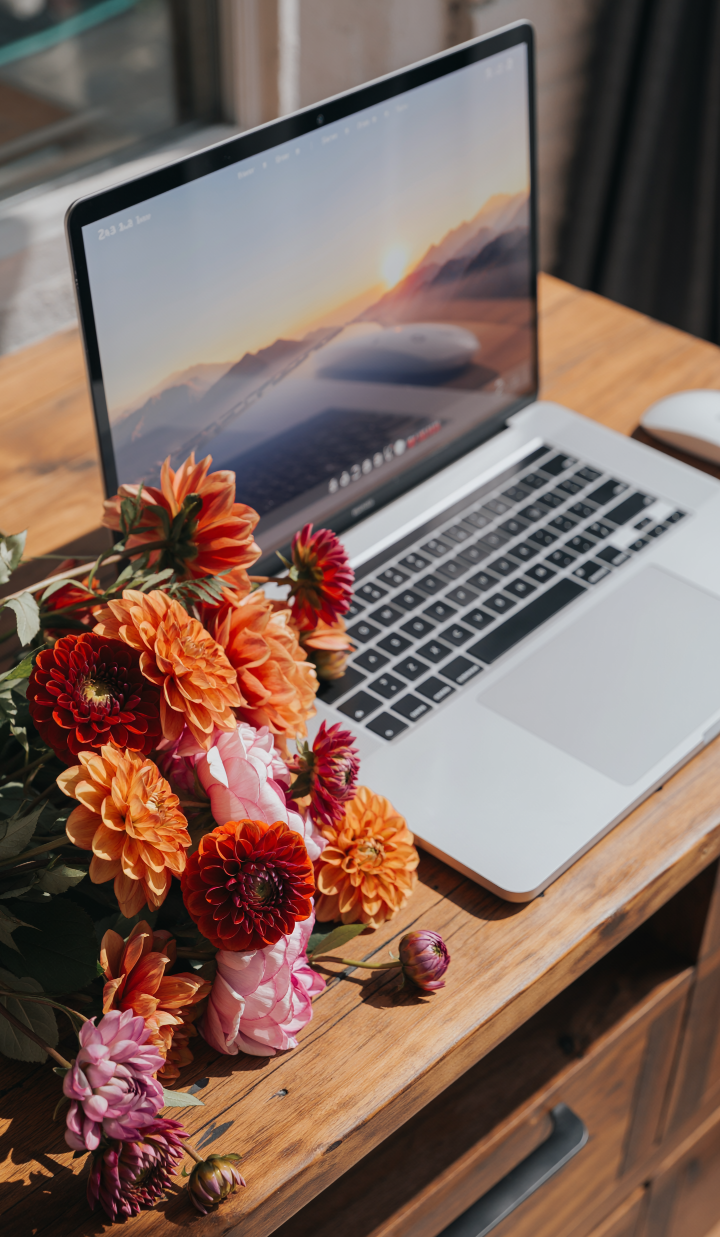 A laptop on a wooden desk with a screen displaying a sunset over mountains and flowers placed next to it.