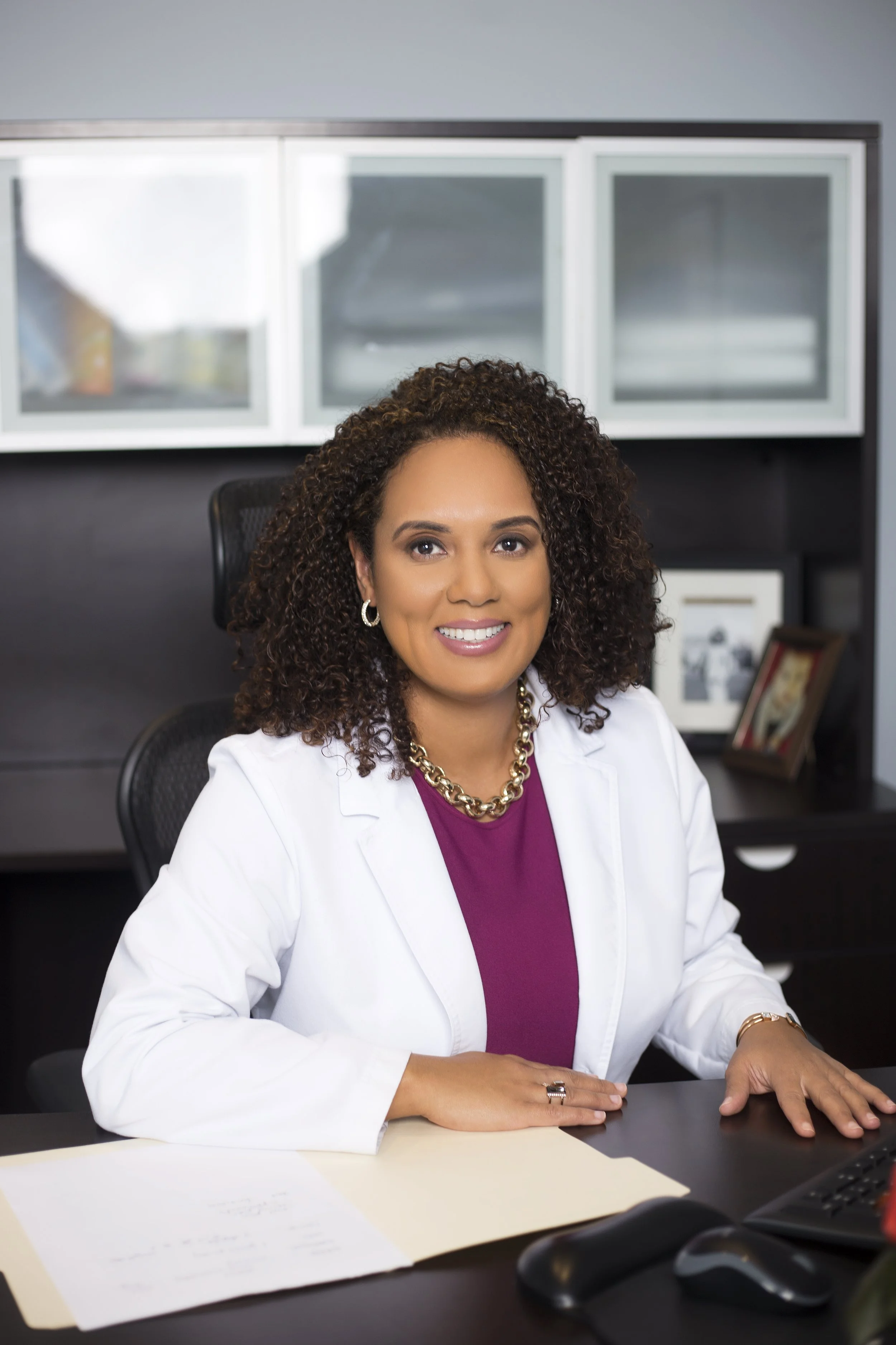 A woman with curly hair wearing a white blazer and a purple top, sitting at a desk in an office.