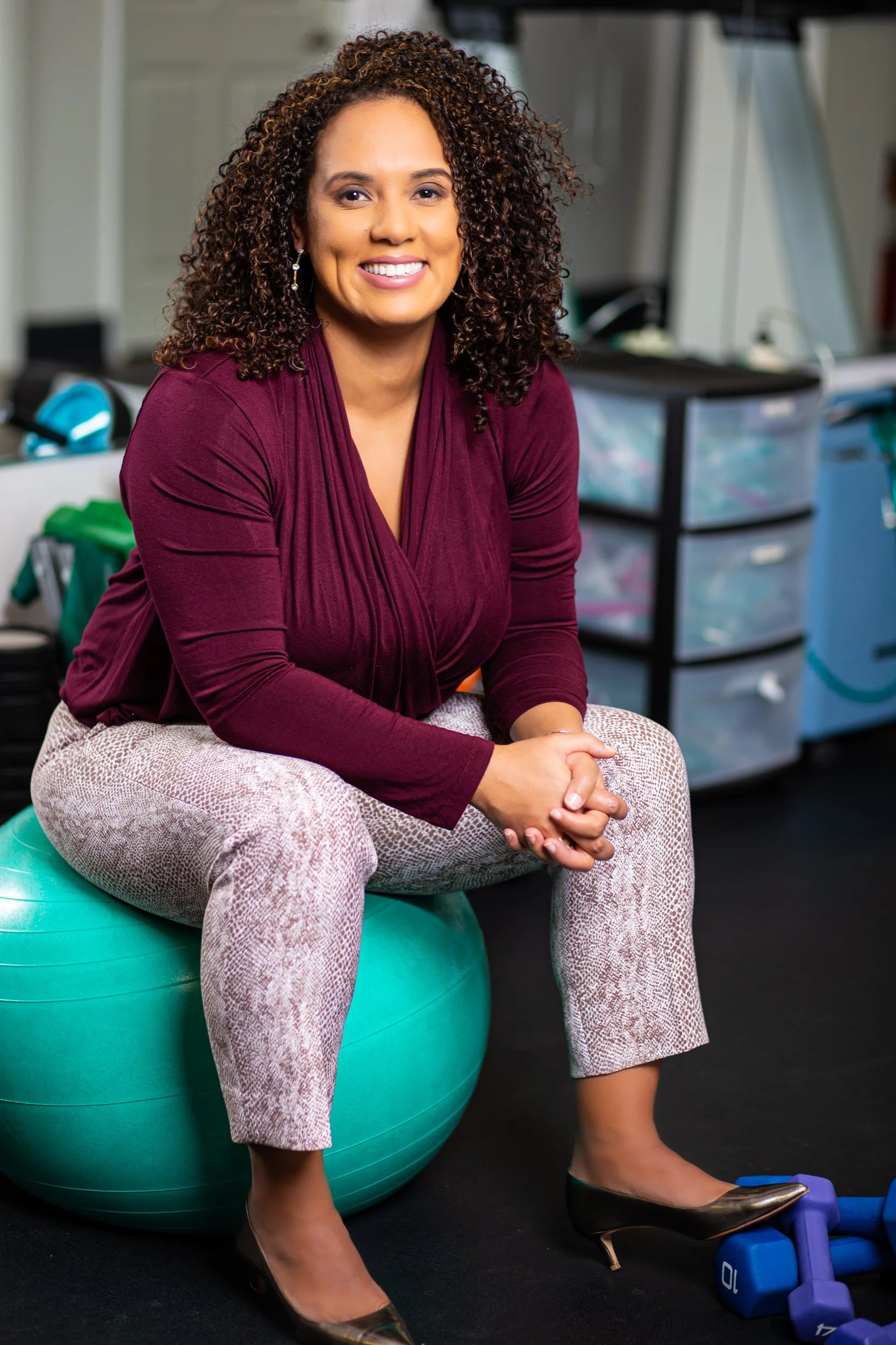A woman with curly hair, smiling, sitting on a teal exercise ball in a fitness or therapy room. She is wearing a maroon top, light patterned pants, and black heels, with gym equipment in the background.