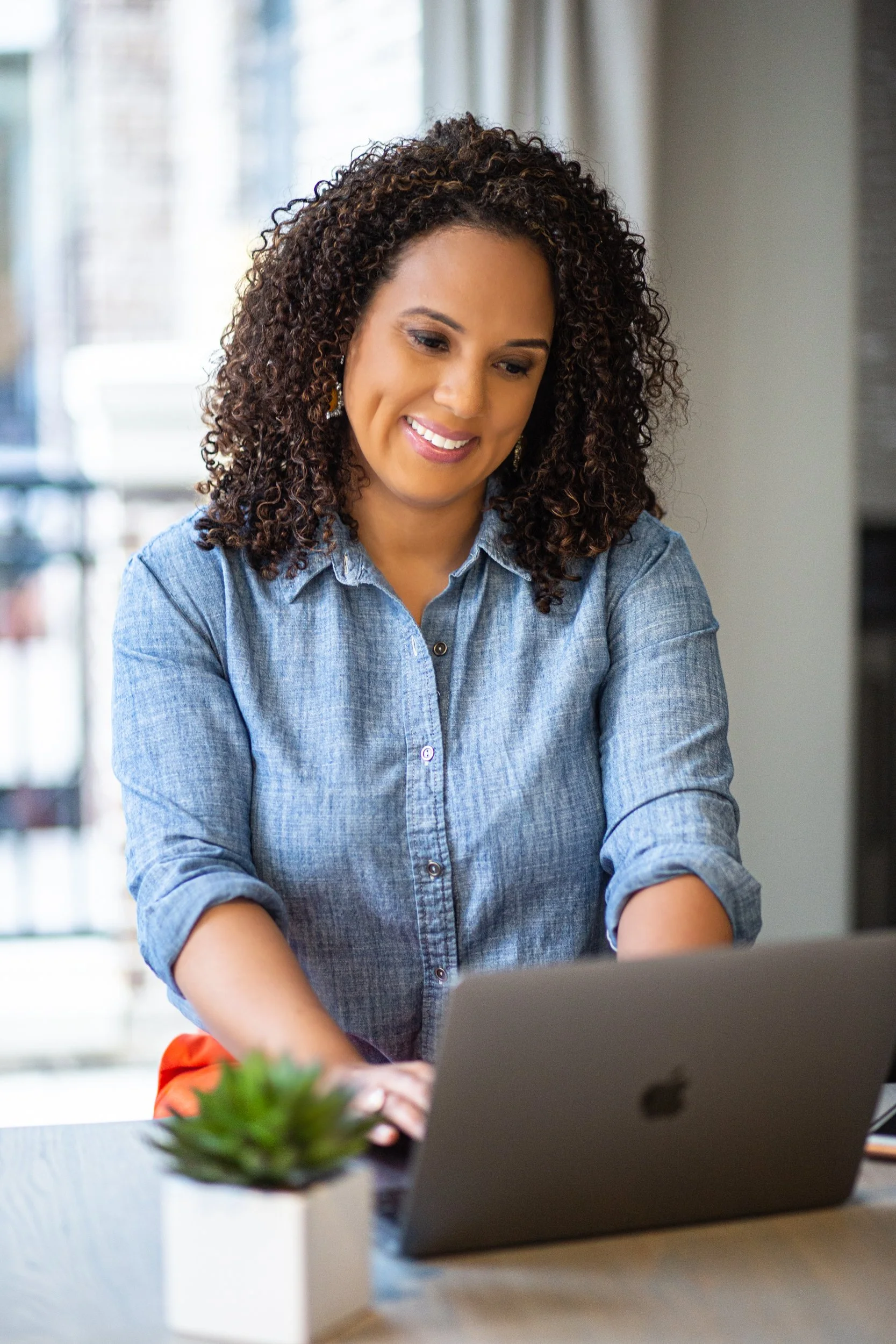 A woman with curly hair is smiling while working on a silver laptop at a desk in a well-lit room.
