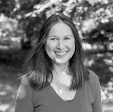 Ein junge Frau mit langem braunem Haar, lachend, im Freien, mit Bäumen im Hintergrund.