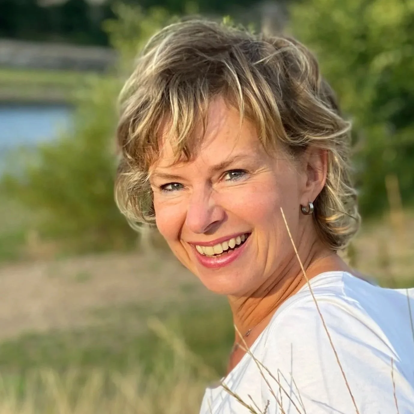 Eine lachende Frau mit kurzen, lockigen, blond-blauen Haaren in einem Outdoor-Umfeld mit Grün und Wasser im Hintergrund.