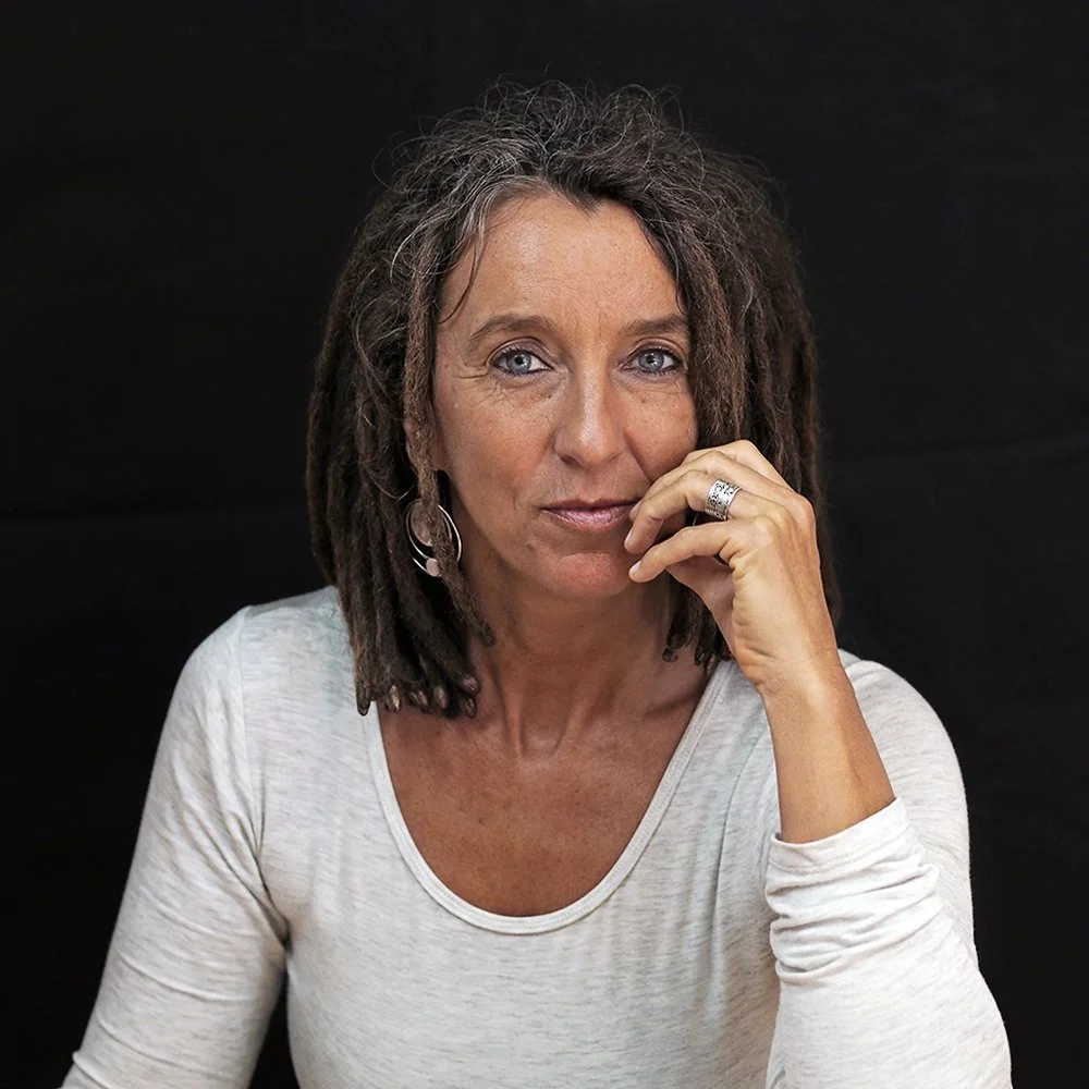 Eine Frau mit lockigem, dunklem Haar und blauer Augen trägt ein hellgraues T-Shirt und betrachtet nachdenklich die Kamera, während sie ihre rechte Hand an ihr Kinn hält.