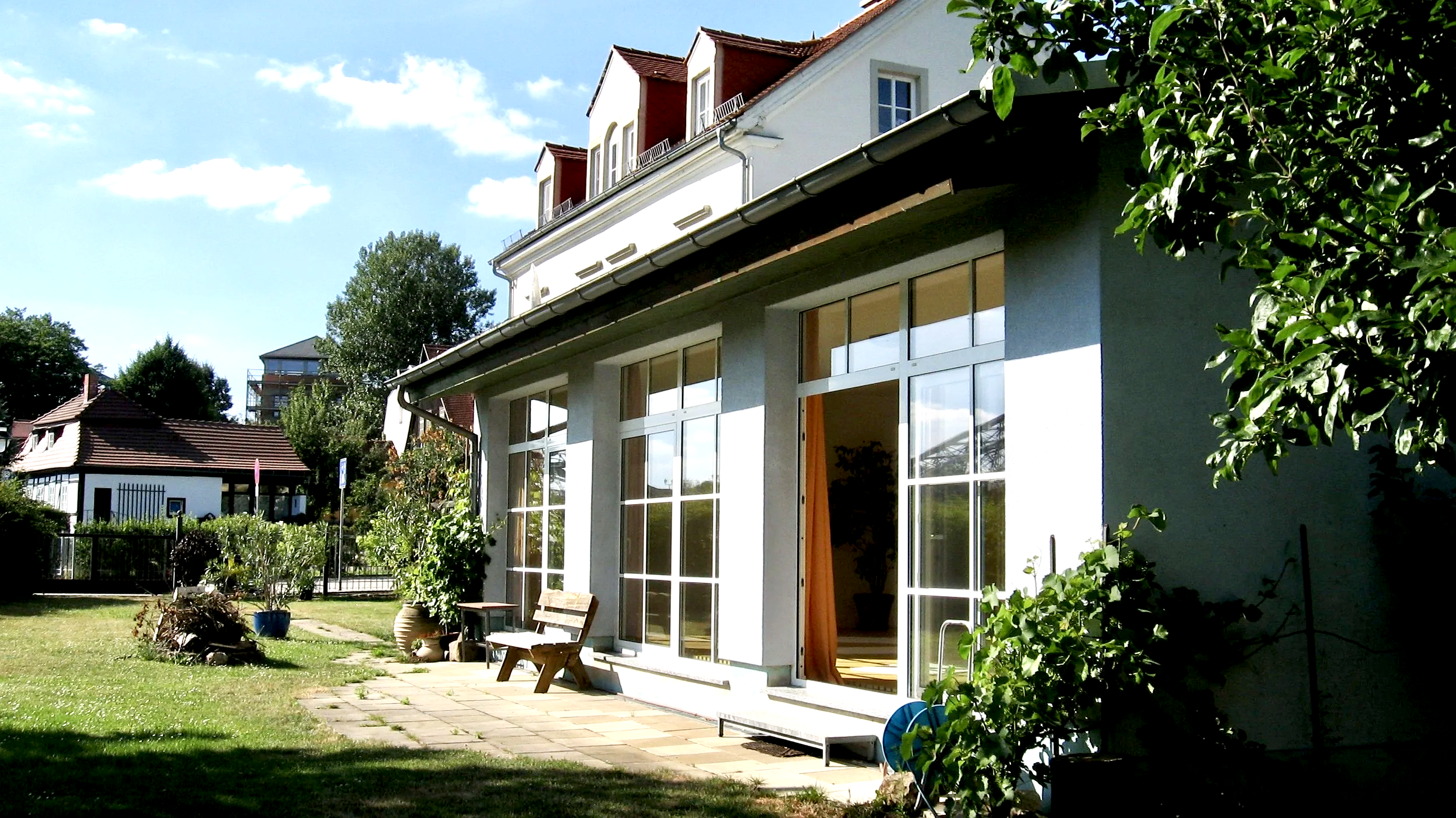 Garten mit Steinterrasse, Bänken, Pflanzen in Töpfen, davor ein Wohnhaus mit großen Fenstern und Gardinen, im Hintergrund andere Häuser und Bäume bei sonnigem Himmel.