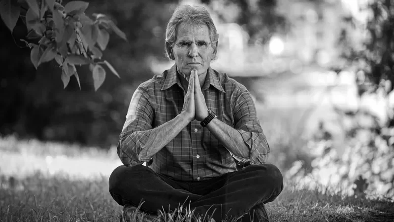 Ein älterer Mann sitzt im Sitzen im Freien auf dem Gras, hält die Hände in Gebetsposition vor seinem Gesicht und sieht ernst.