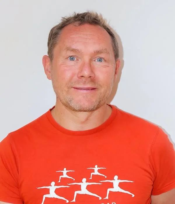 Ein Mann mit blauen Augen und kurzen, hellen Haaren trägt ein rotes T-Shirt mit weißen Yogabildern.